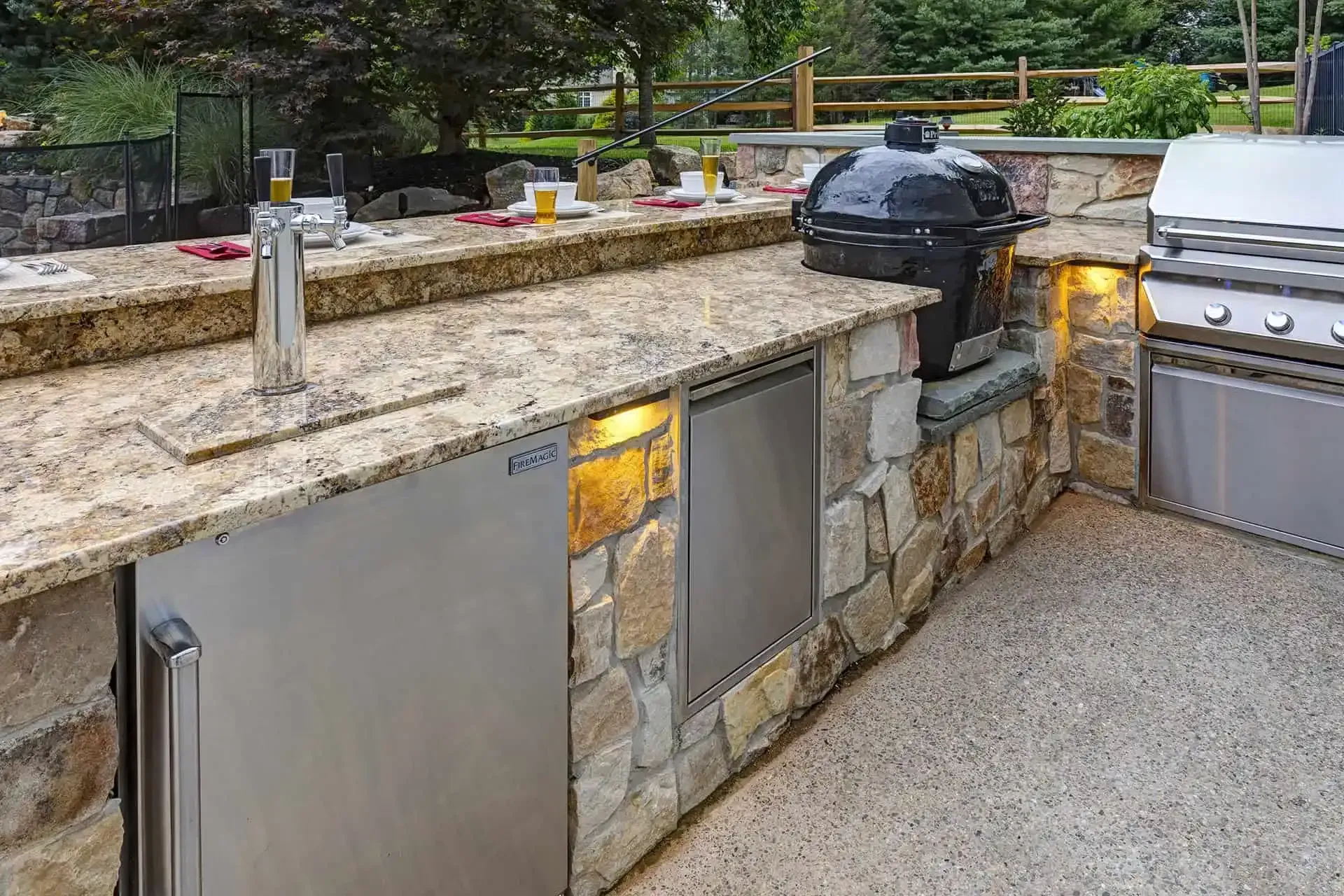 Outdoor stone kitchen with granite counter, built-in grill, black smoker, and stainless steel storage doors