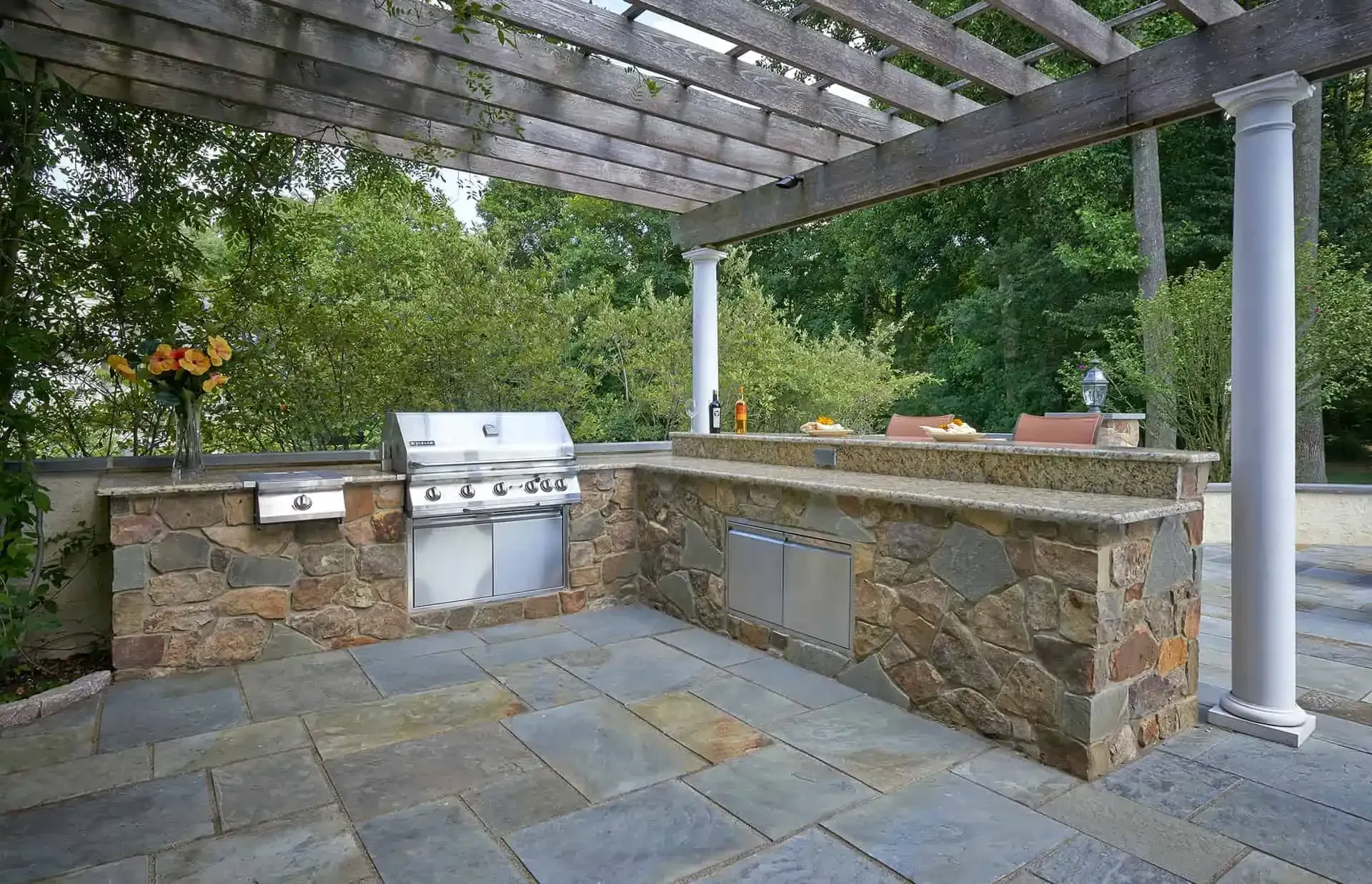 Stone outdoor kitchen with grill under a pergola, surrounded by greenery
