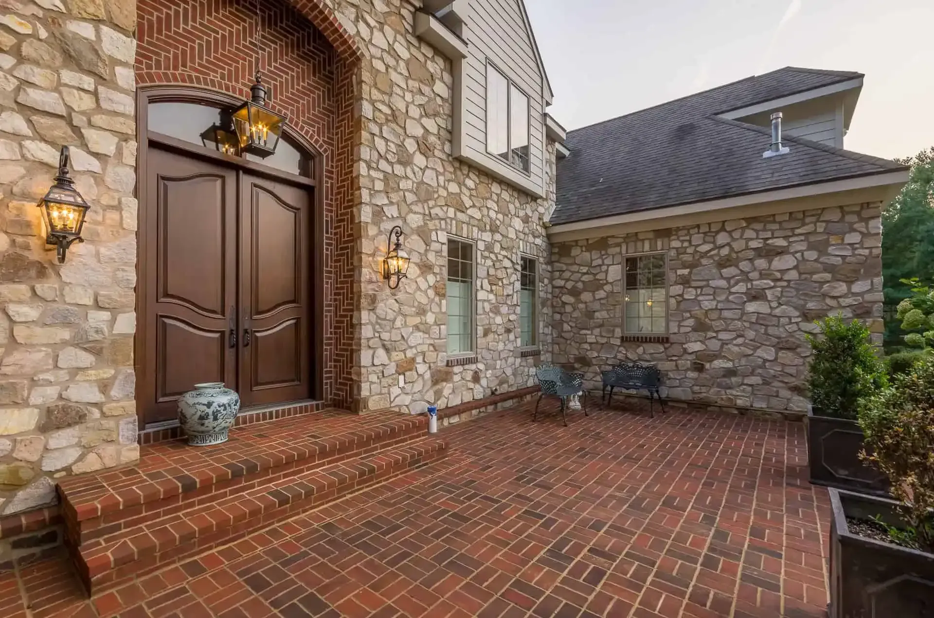 Stone front entry with wooden double doors and red brick patio beside a house