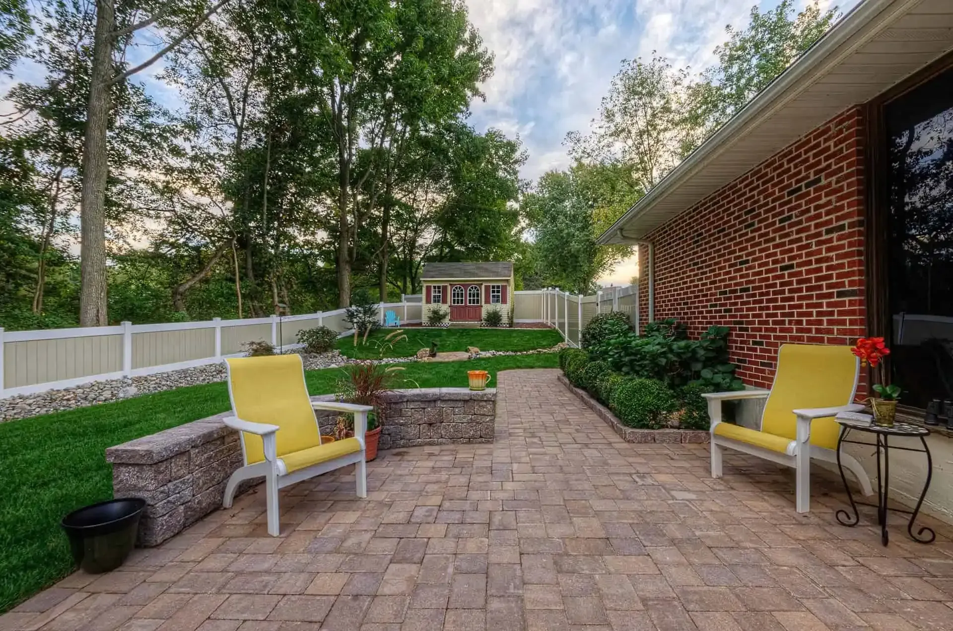 Brick patio with yellow chairs, raised garden beds, and a fenced backyard beside a brick house