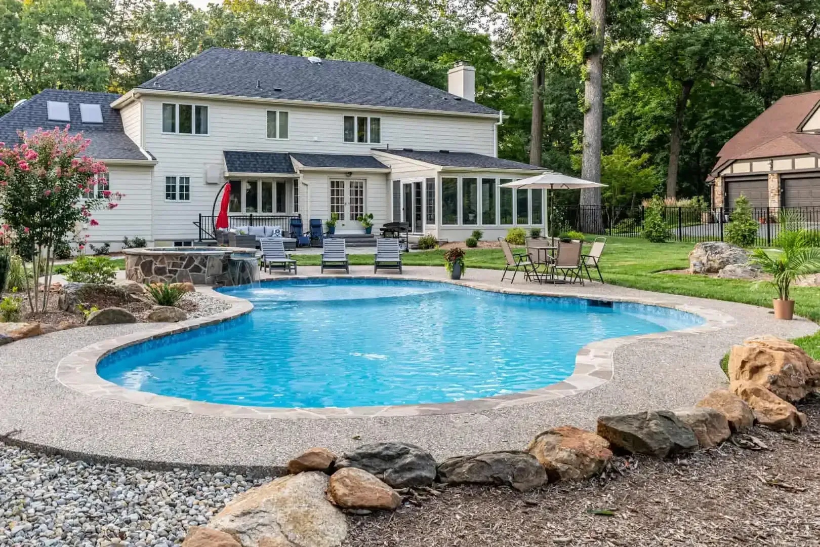 Backyard with a blue swimming pool, stone edging, patio seating, and a white house surrounded by trees.