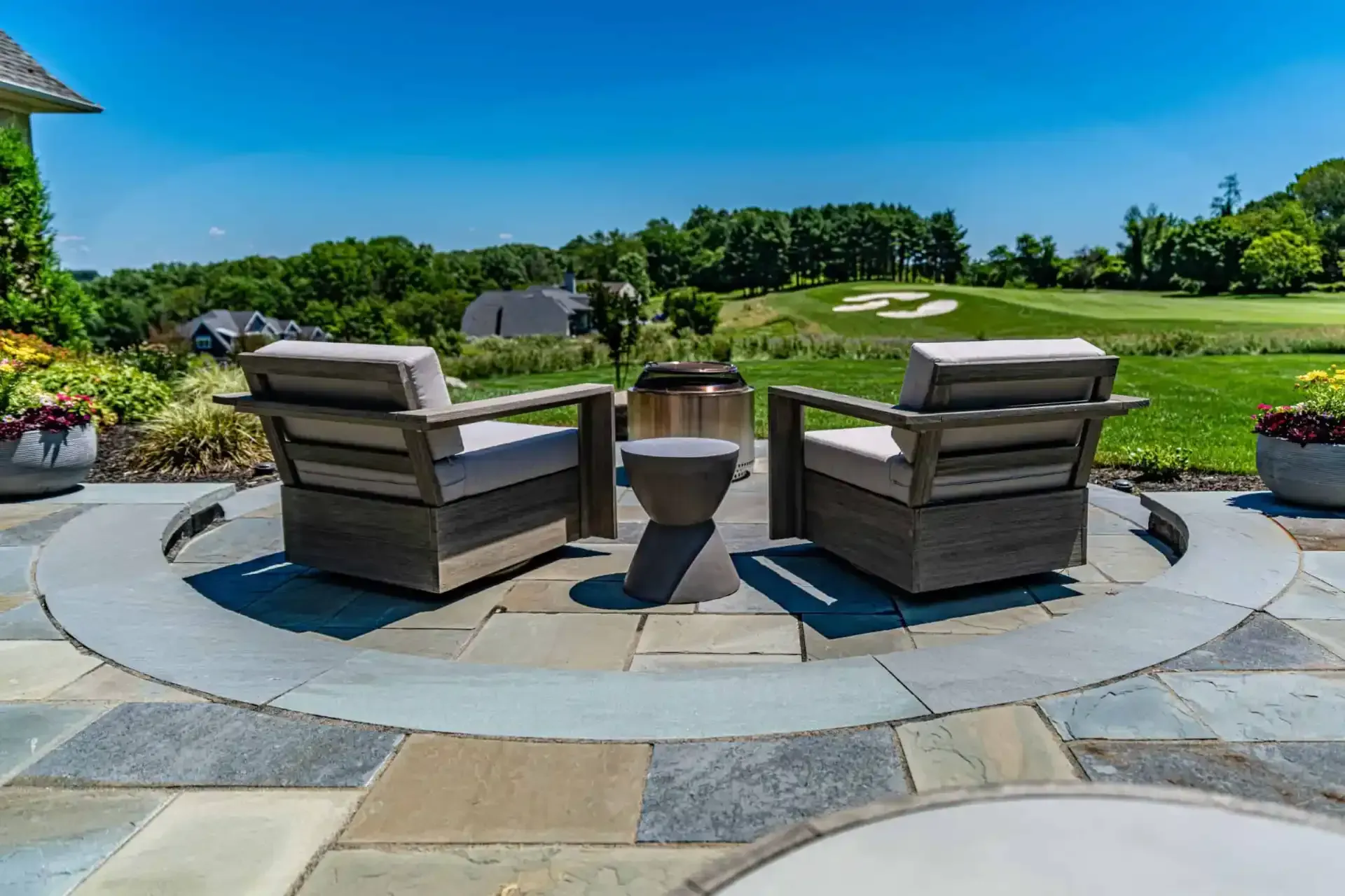 Two gray Adirondack chairs facing a fire pit on a stone patio overlooking a green lawn and golf course