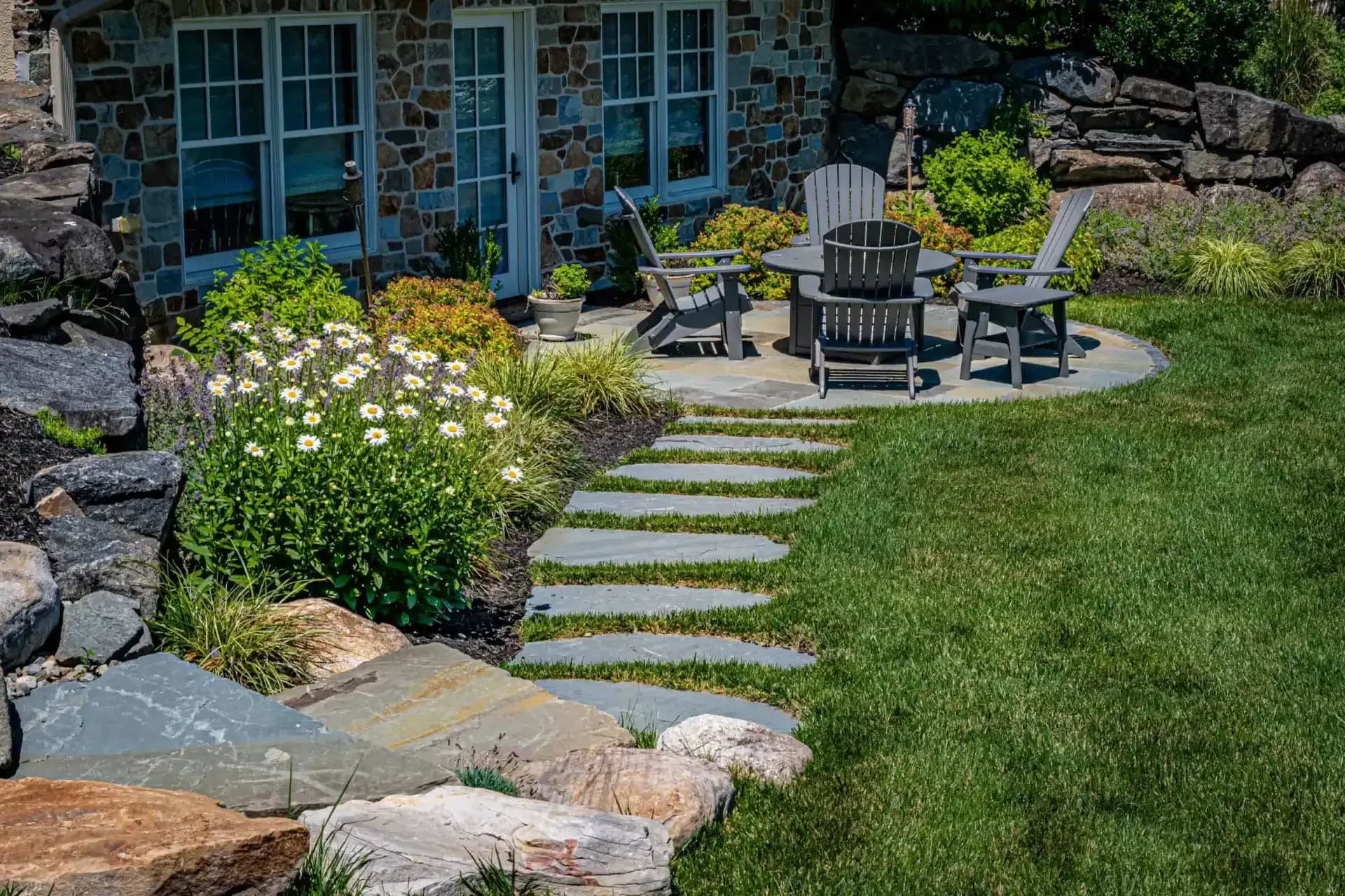 Stone steps through a garden lead to a blue house patio with black chairs and white flowers