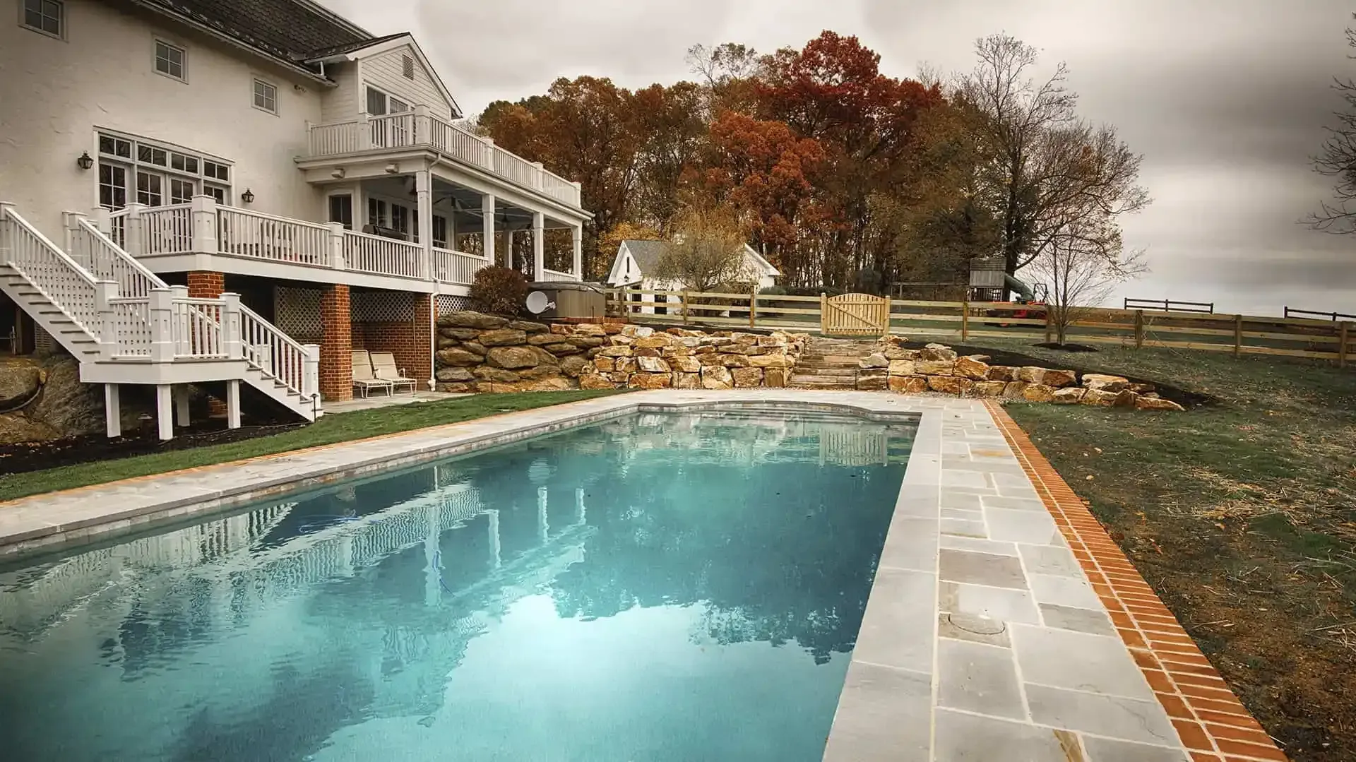 Backyard swimming pool beside a white house, with autumn trees and a cloudy sky.