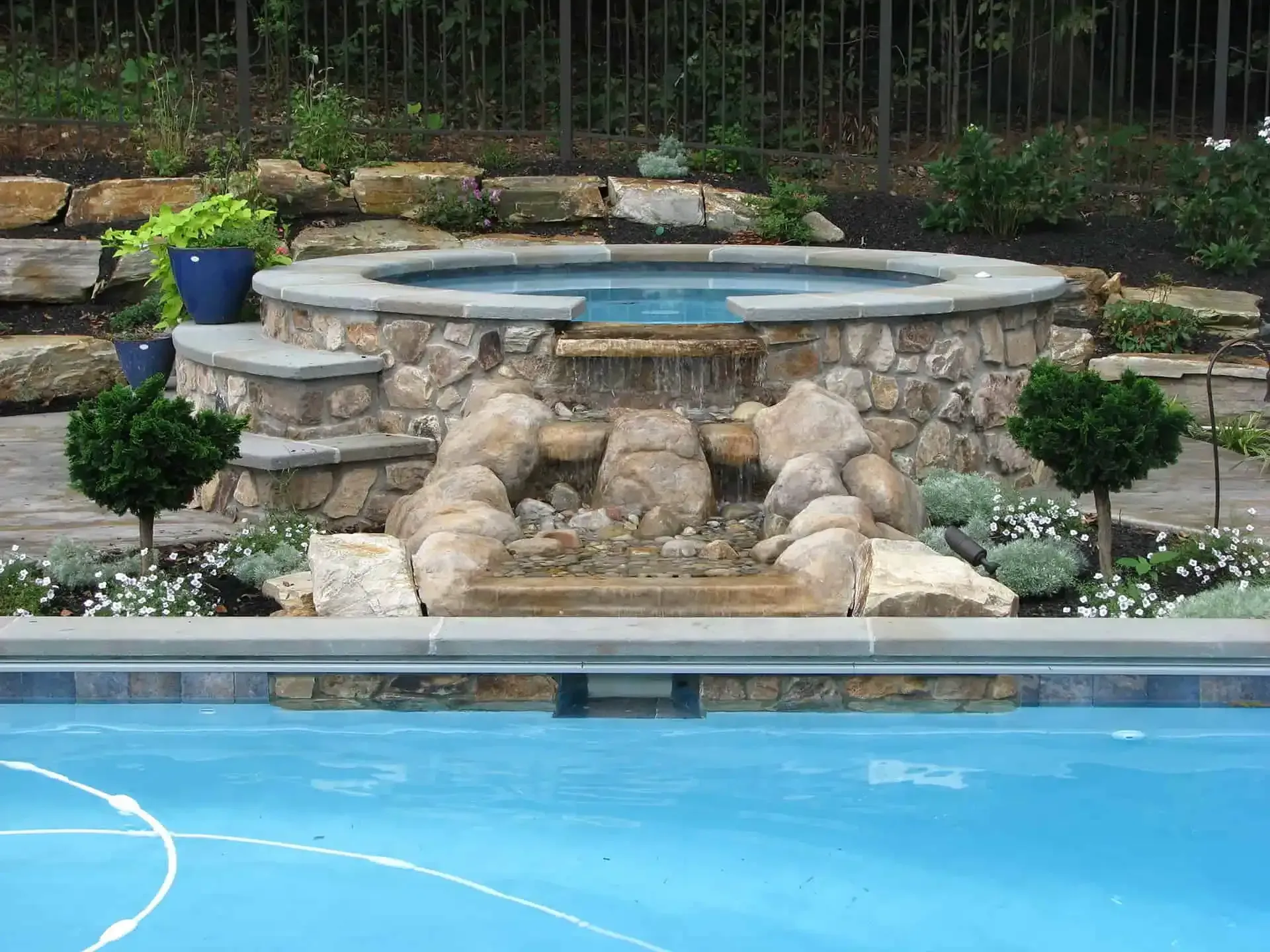 Stone hot tub beside a bright blue swimming pool with landscaped rocks and shrubs