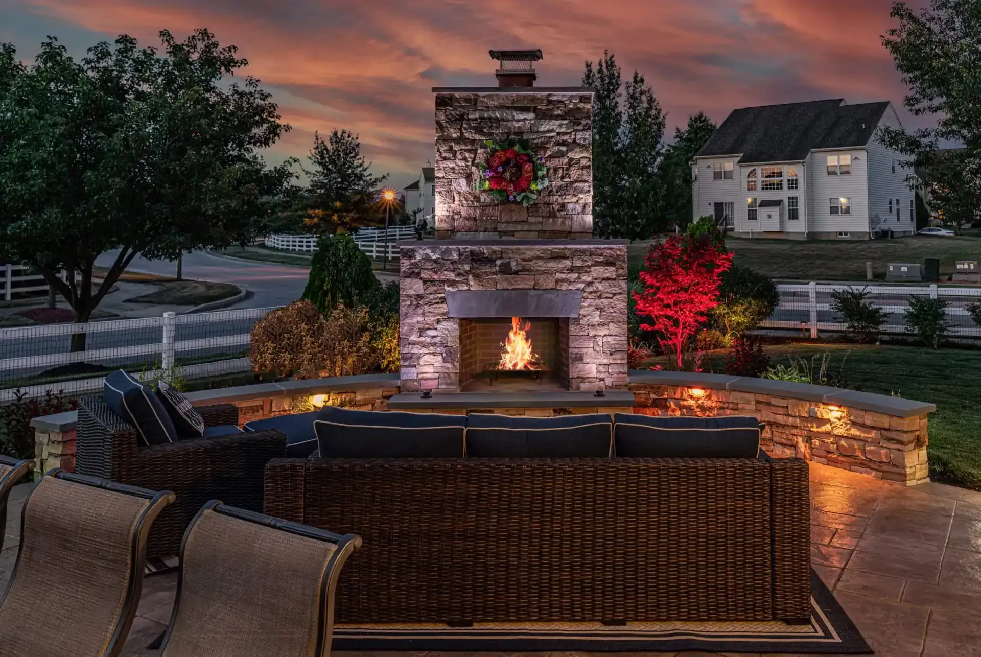 Backyard fire pit and stone fireplace at dusk with wicker seating and glowing landscape lights