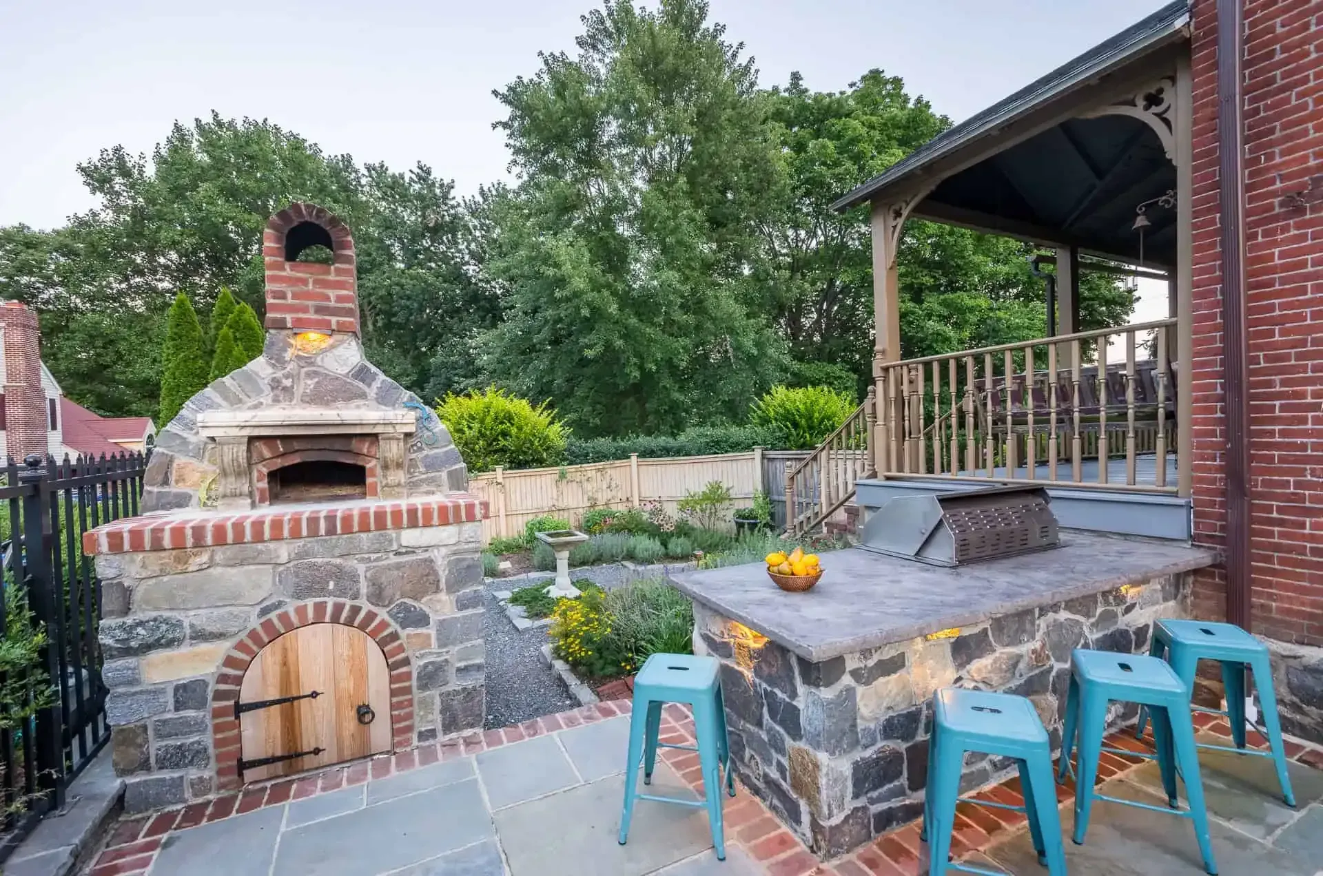 Stone outdoor patio with brick pizza oven, grill, and blue stools beside a wooden porch.
