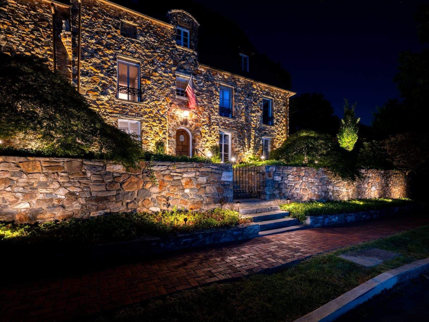 Stone building and garden wall lit at night, with a lit doorway and steps beside a brick path