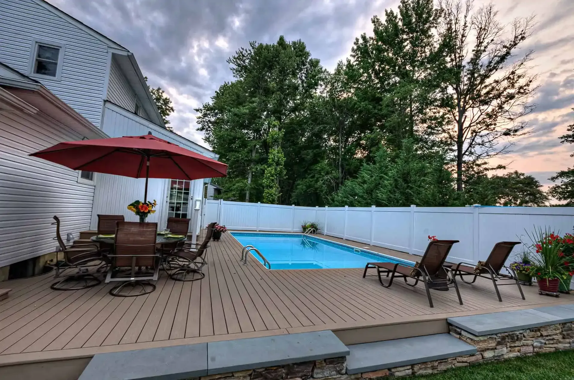 Backyard patio with pool, lounge chairs, red umbrella, and evening sky beside a house