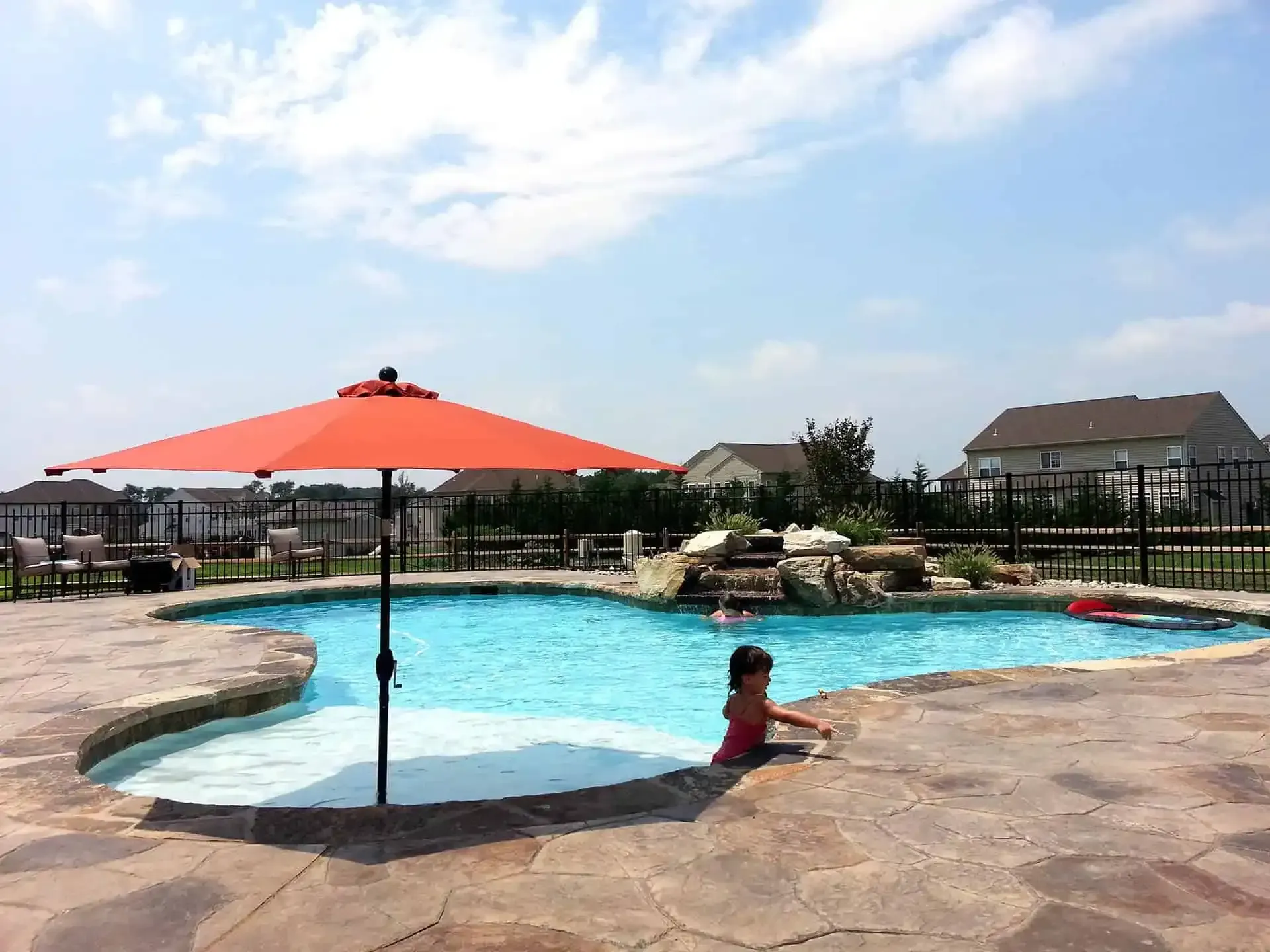 Resort pool with orange umbrella, patio deck, and a child sitting at the water’s edge under a blue sky