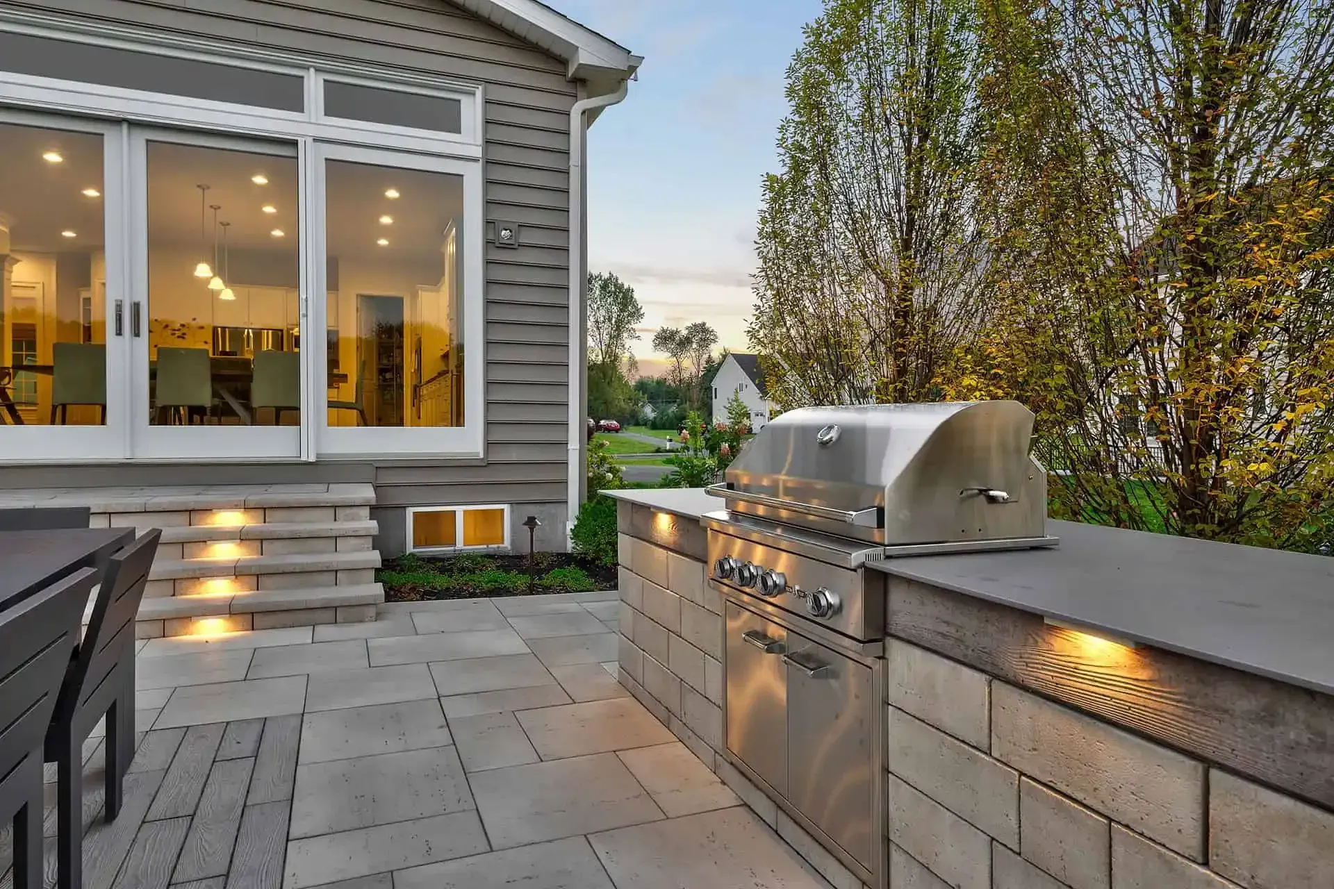 Outdoor patio beside a house with stone pavers, built-in grill, and warm lights at dusk
