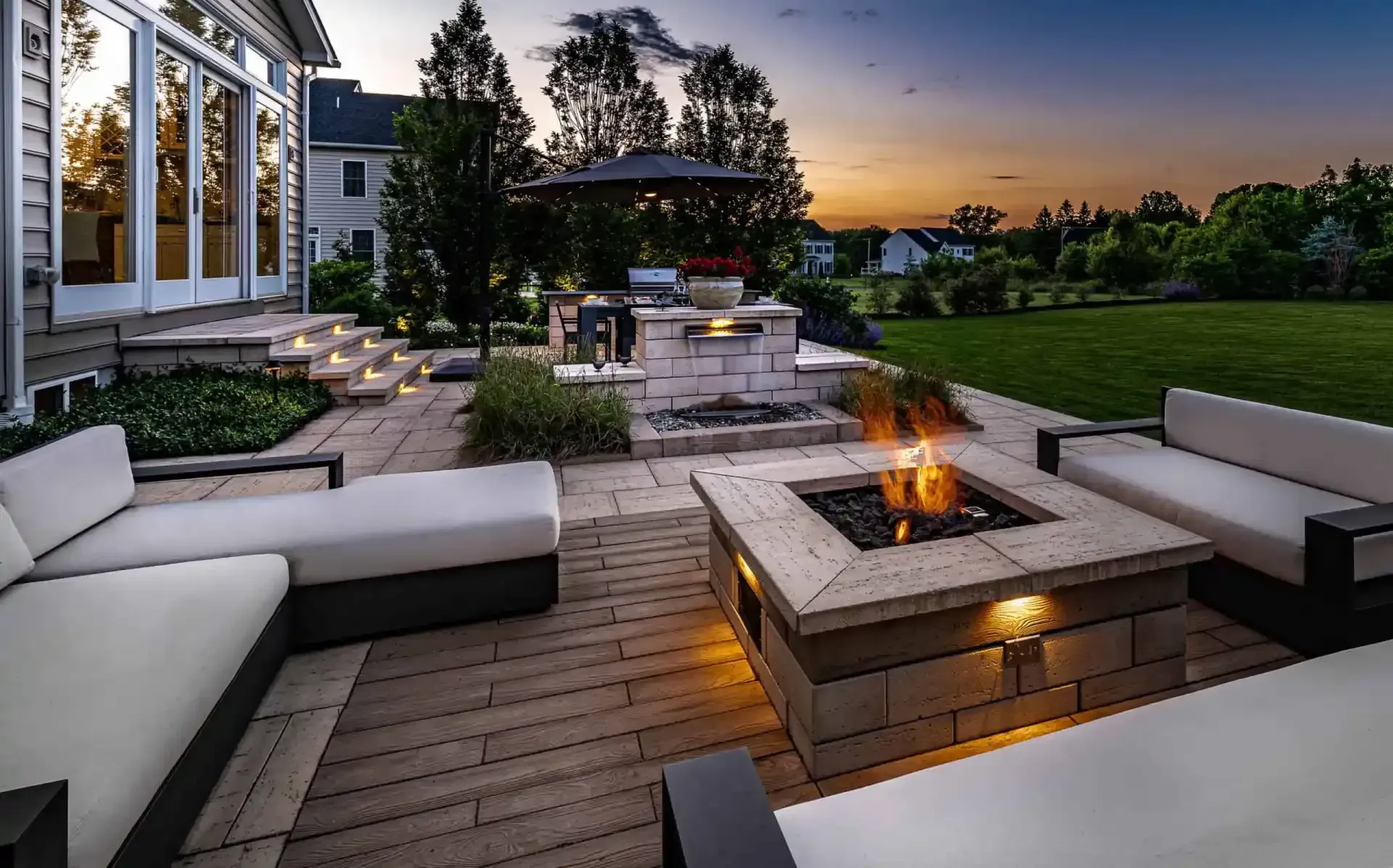Modern backyard patio with white sofas, stone fire pit, and sunset view over green lawn