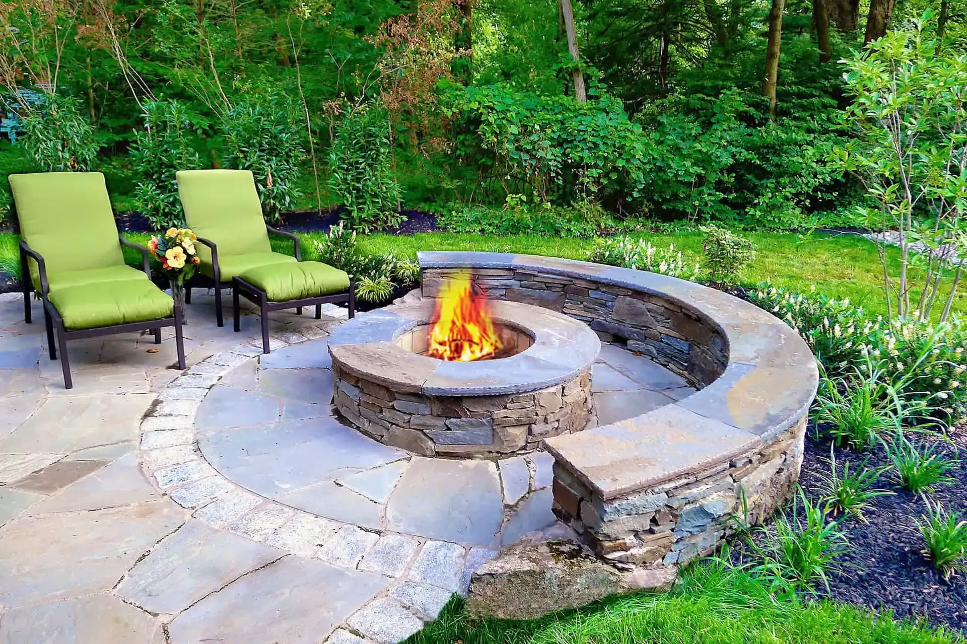 Backyard stone fire pit with curved bench, two green lounge chairs, and a wooded garden backdrop