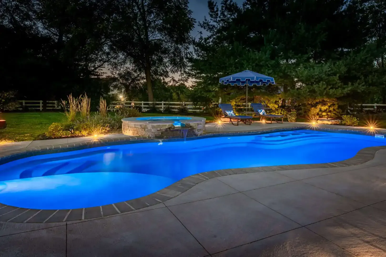 Blue-lit backyard swimming pool at dusk with patio seating and garden lights