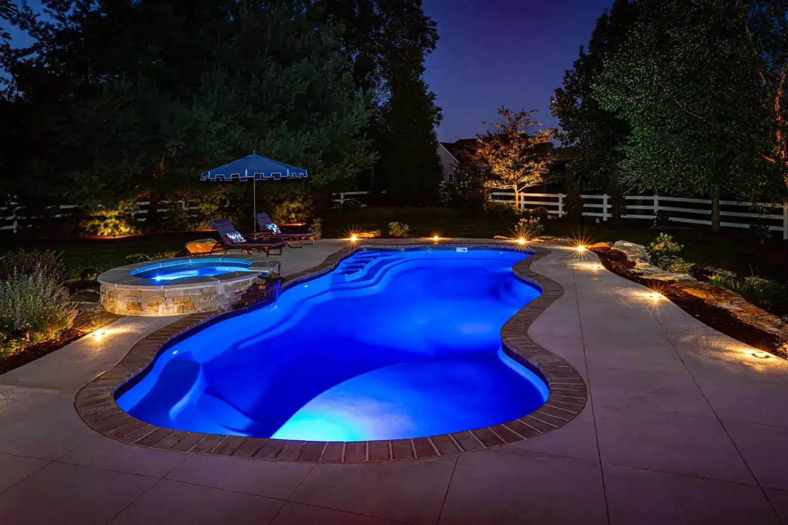 Blue-lit backyard swimming pool at night with patio, hot tub, and surrounding trees