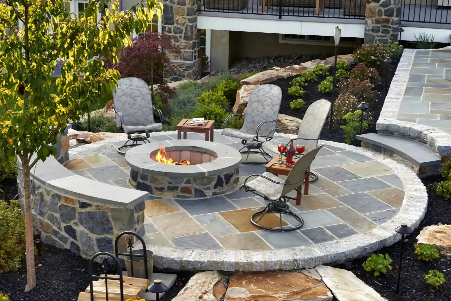 Stone patio fire pit area with curved seating, Adirondack chairs, and landscaped garden steps