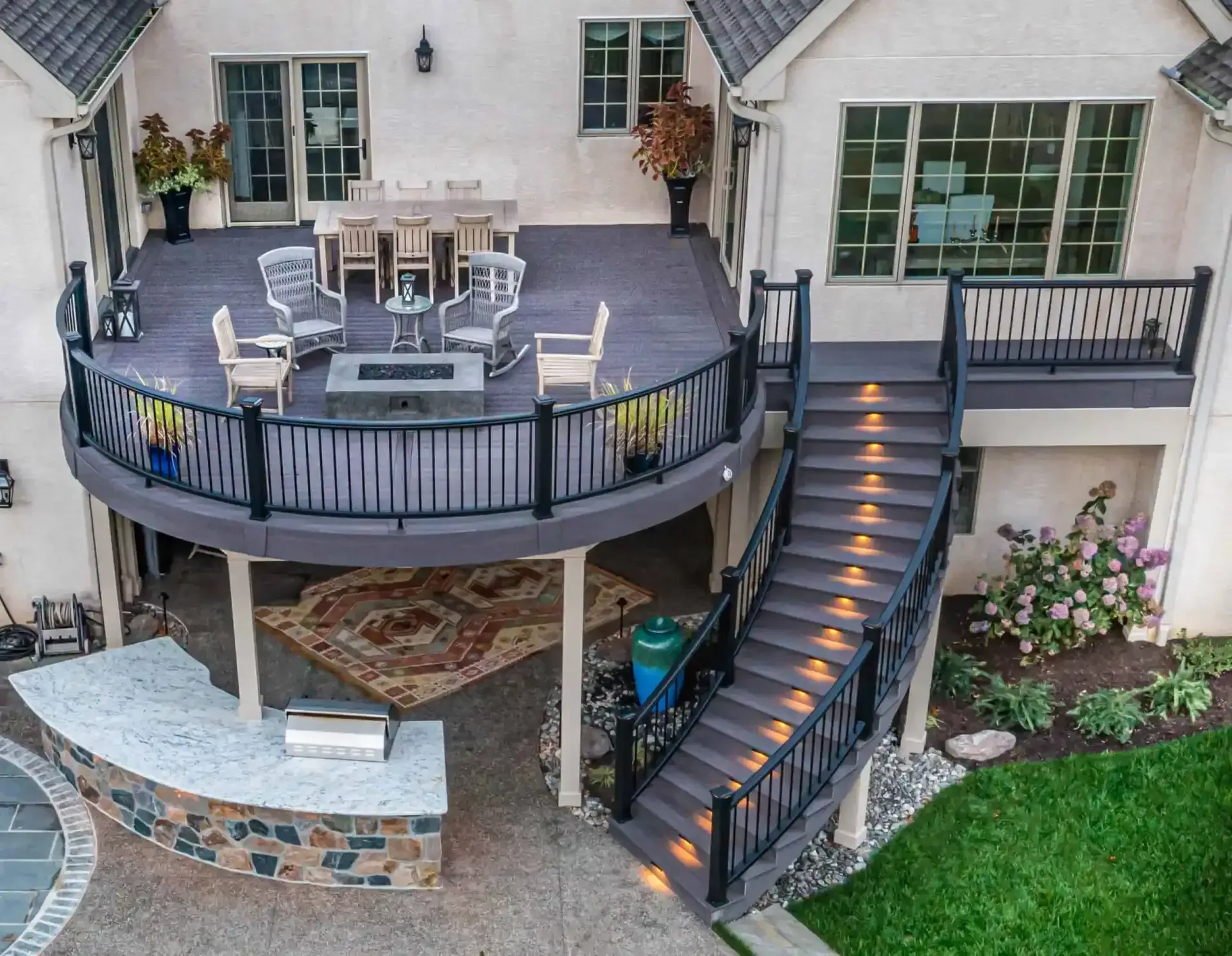 Backyard patio with curved deck, outdoor dining set, lit stairs, and stone seating area