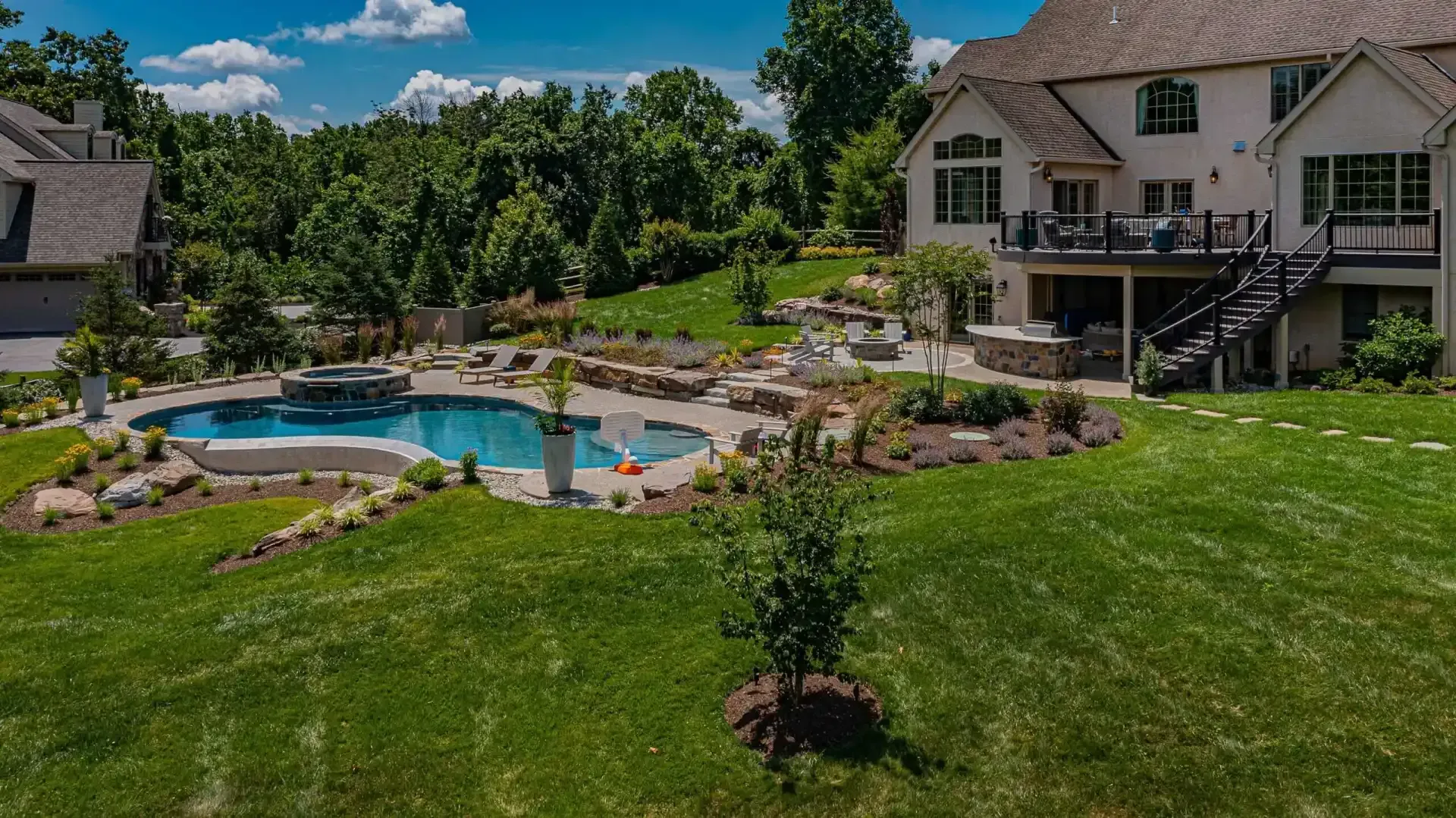 Backyard with a swimming pool, stone patio, and large house overlooking a green lawn and trees.