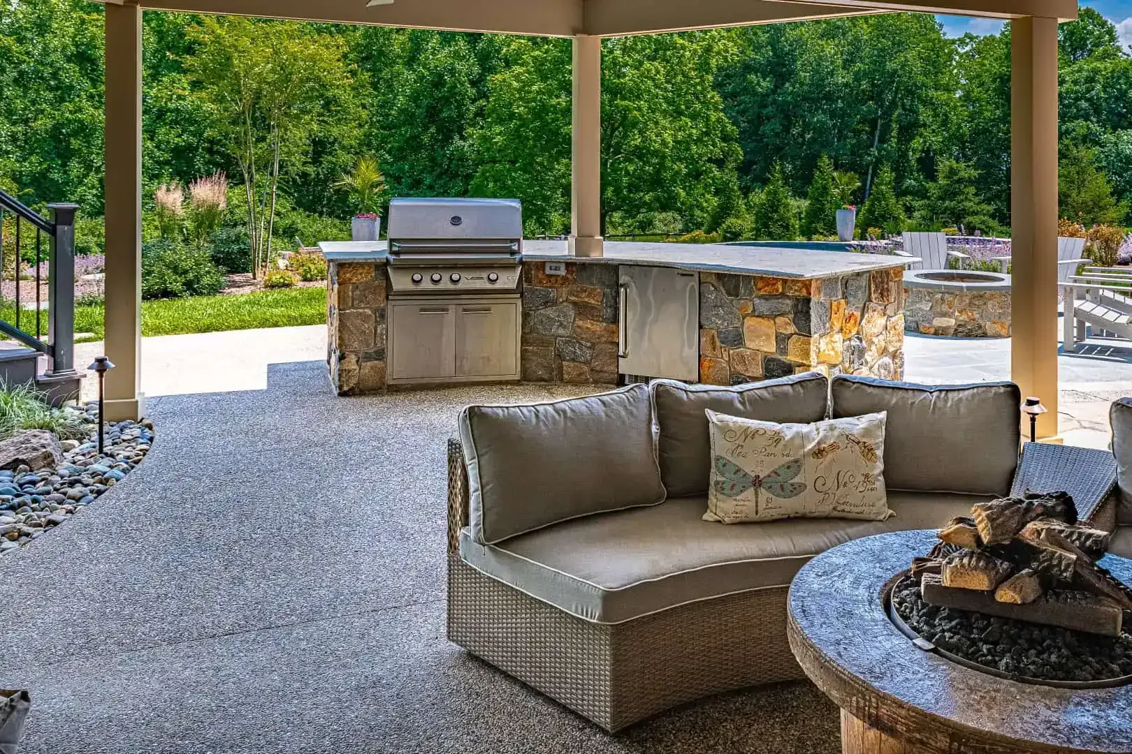 Covered patio with outdoor sofa, fire pit, grill, and stone bar overlooking a landscaped yard