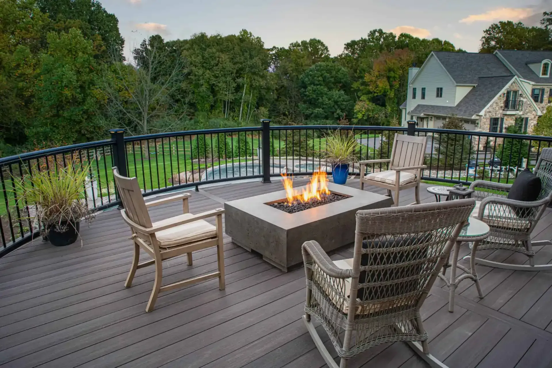 Back deck with wicker chairs around a lit fire pit, overlooking trees and a house at dusk