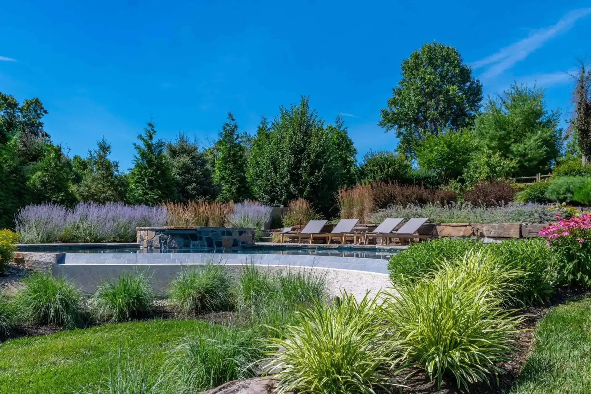 Landscaped poolside patio with lounge chairs, lush greenery, and a clear blue sky