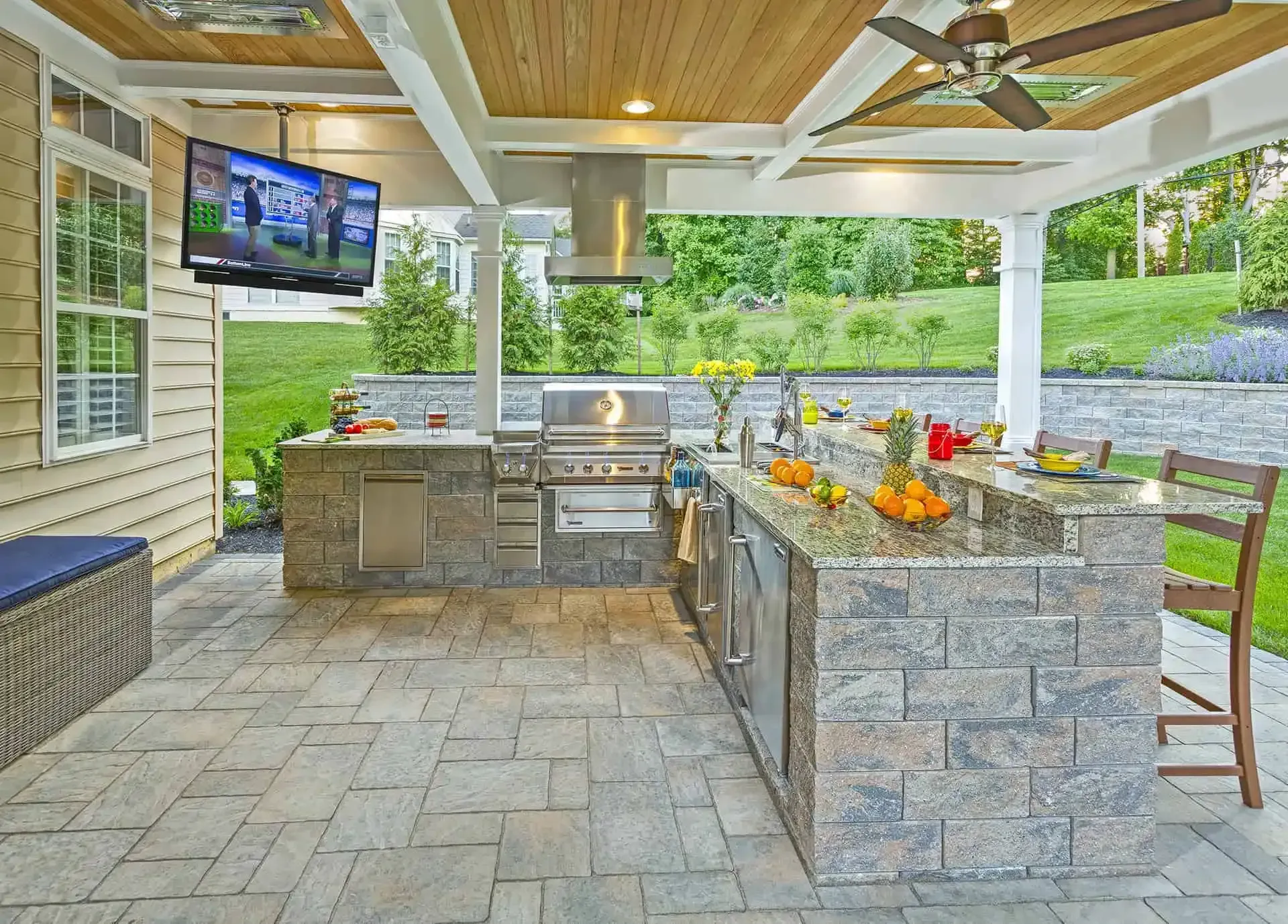 Covered outdoor kitchen and patio with stone counters, grill, bar stools, TV, and potted flowers