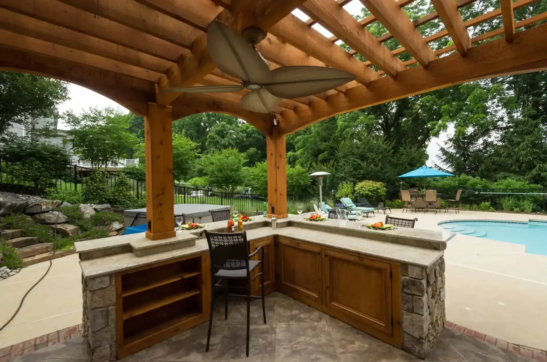 Outdoor poolside bar under a wooden pergola with stone counters, stools, and greenery nearby