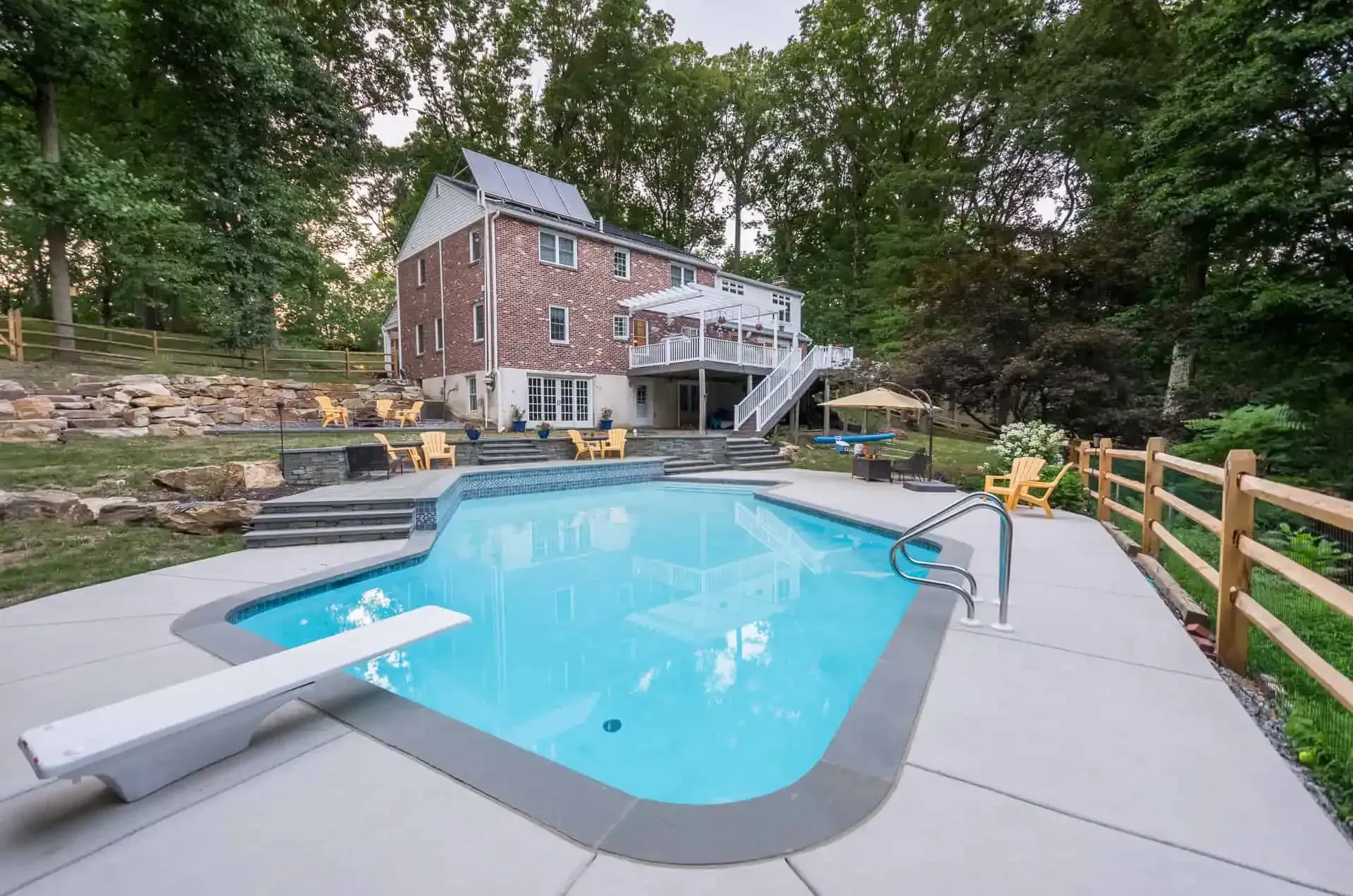 Backyard pool with diving board, stone patio, and brick house surrounded by trees