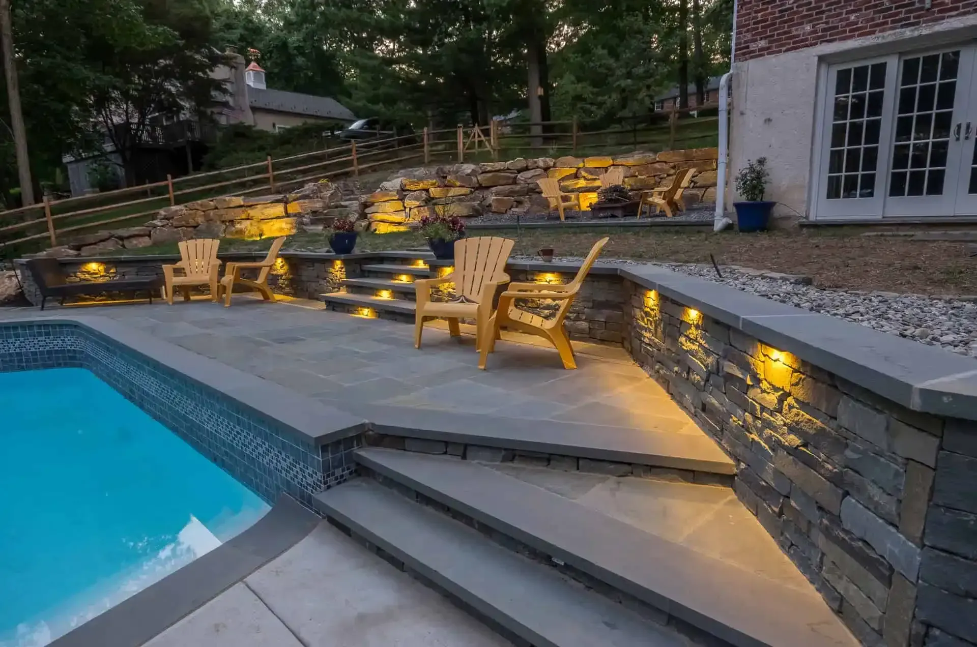 Backyard pool at dusk with lit stone seating terraces and patio chairs