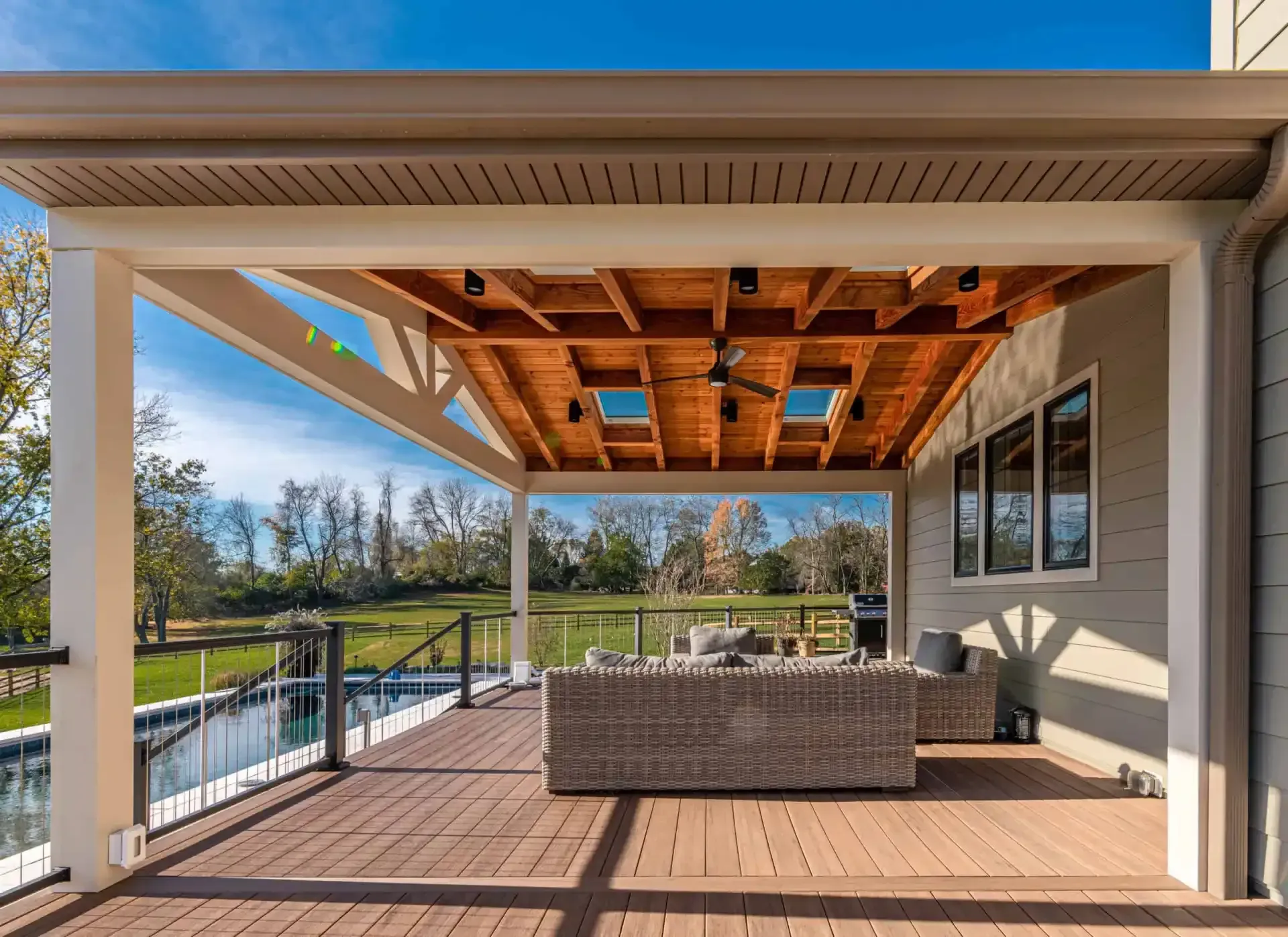 Covered backyard patio with wicker seating, wood ceiling, and a view of trees and water beyond the railing
