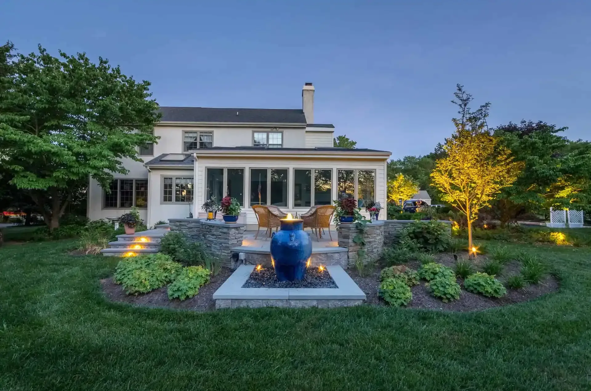 Suburban house at dusk with lit garden, stone patio, and fountain in the foreground