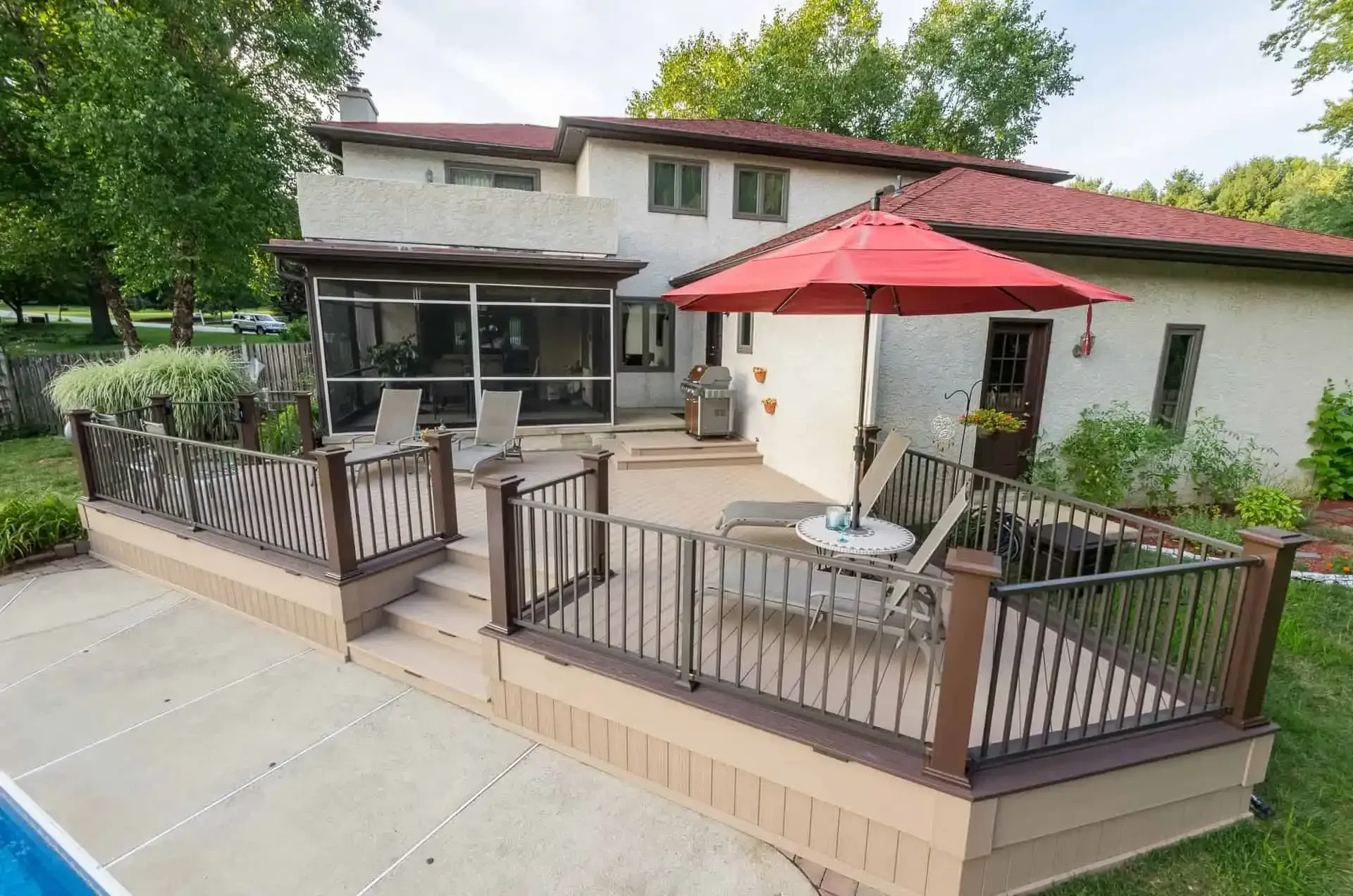 House with red-roofed patio deck, railing, lounge chairs, and red umbrella in a backyard.