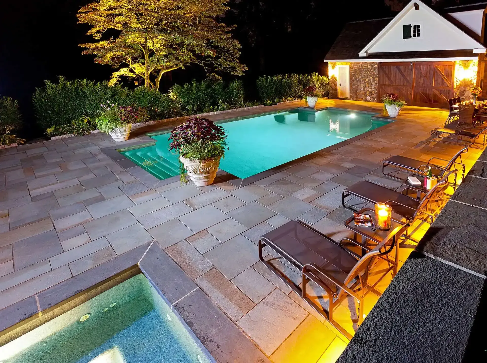 Backyard pool and hot tub at night with glowing lights, lounge chairs, and a garage in the background