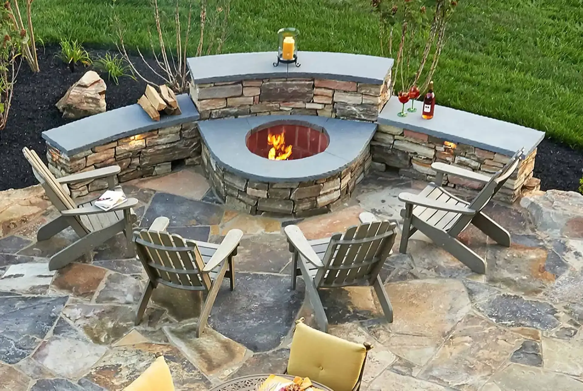 Stone patio fire pit with Adirondack chairs arranged around a circular seating area outdoors.