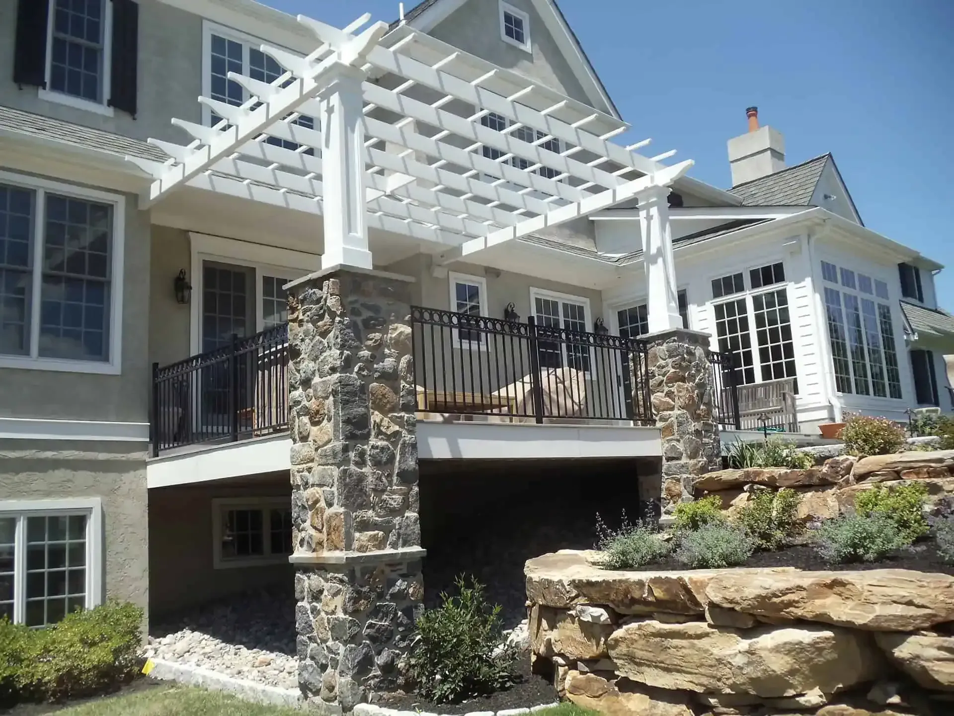 Stone-and-wood house exterior with a white pergola over a raised deck and stone retaining wall.