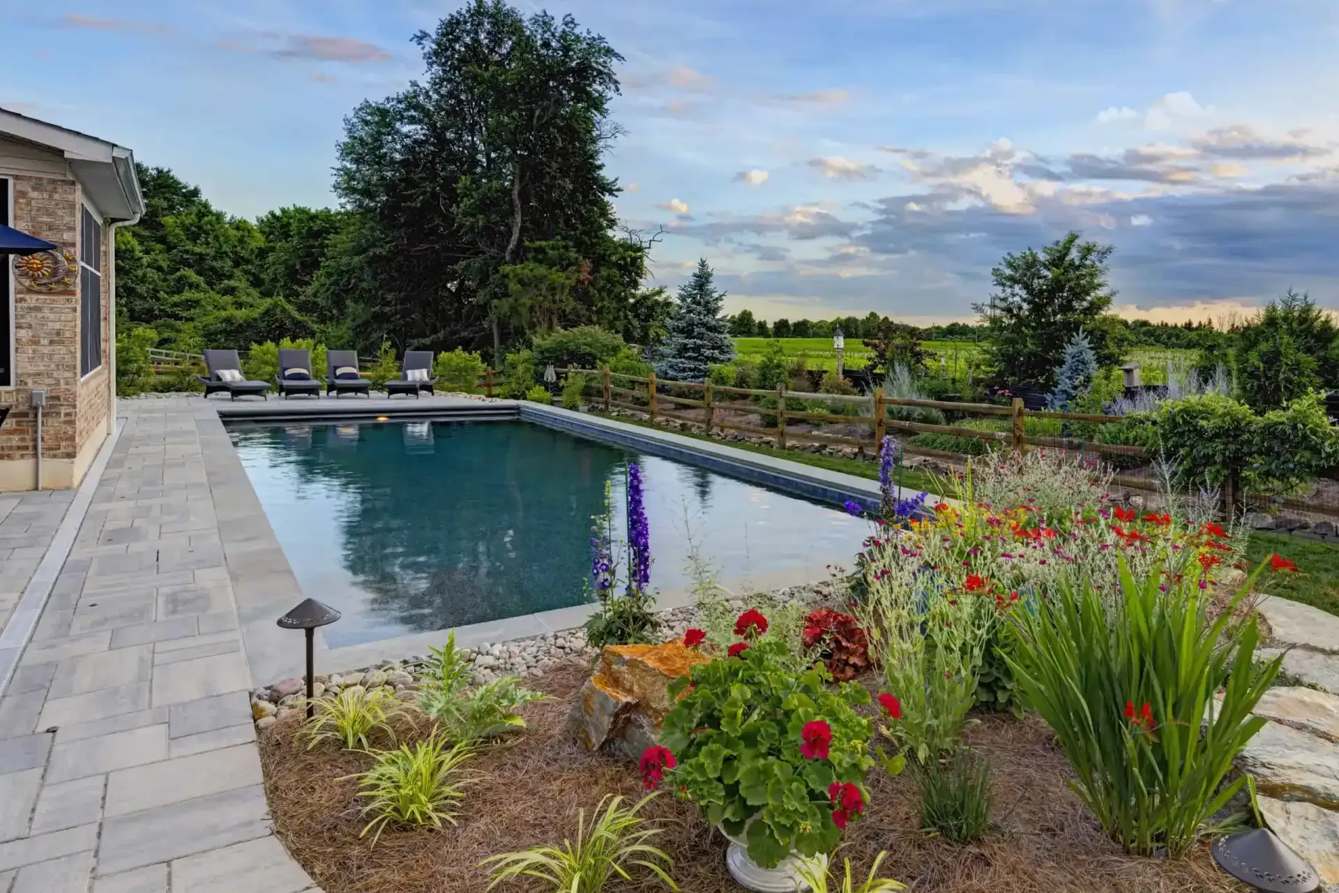 Backyard pool with stone patio, lounge chairs, colorful flowers, and landscaped garden under a cloudy sky