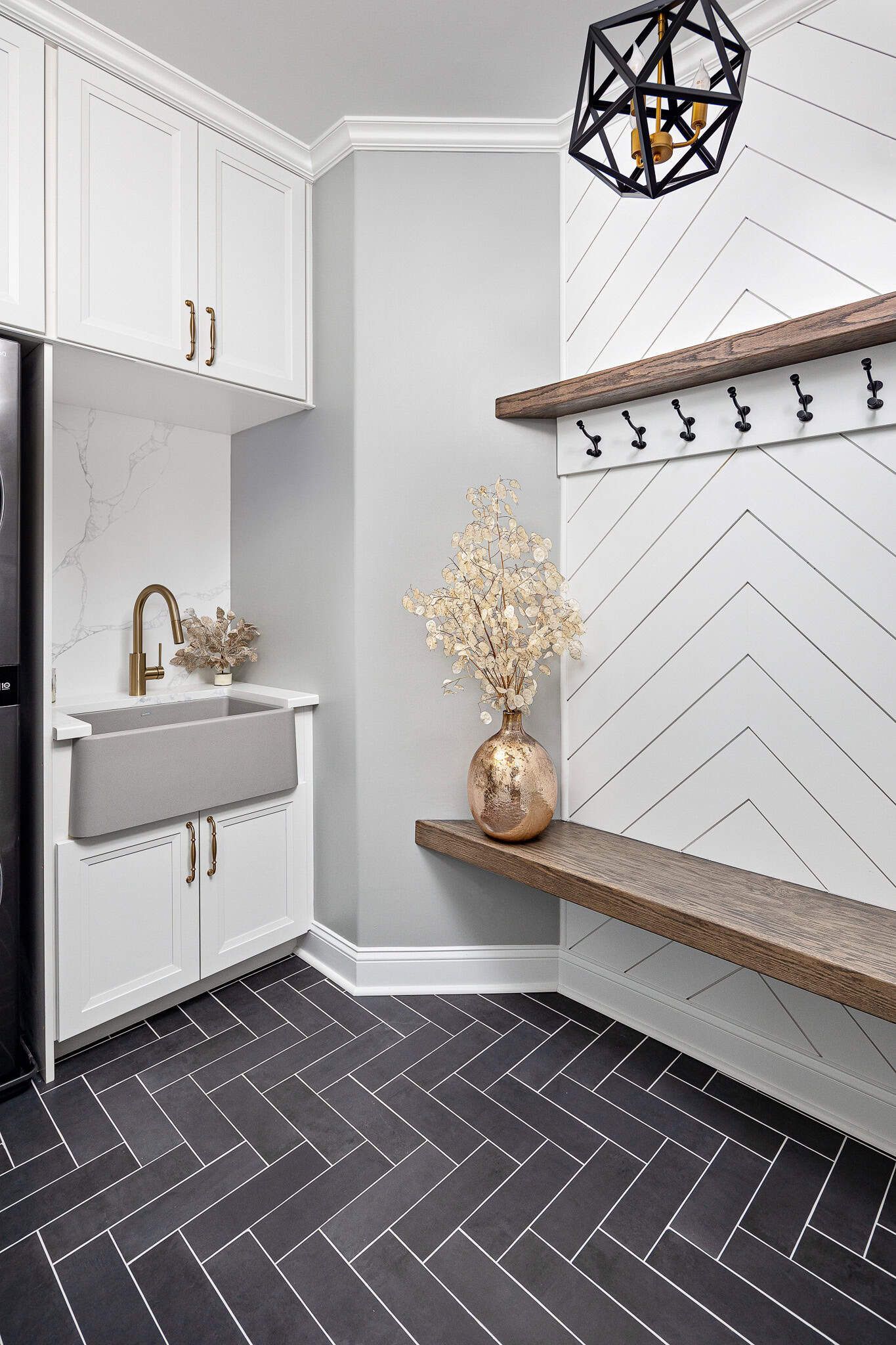 Modern mudroom with white cabinets, small sink, wood bench, hooks, geometric pendant light, and dark herringbone floor.