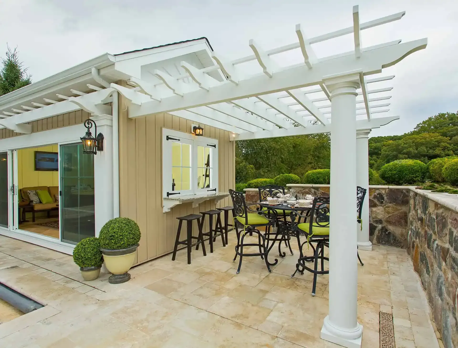 Covered patio with white pergola, bistro table and chairs beside a beige house and stone wall