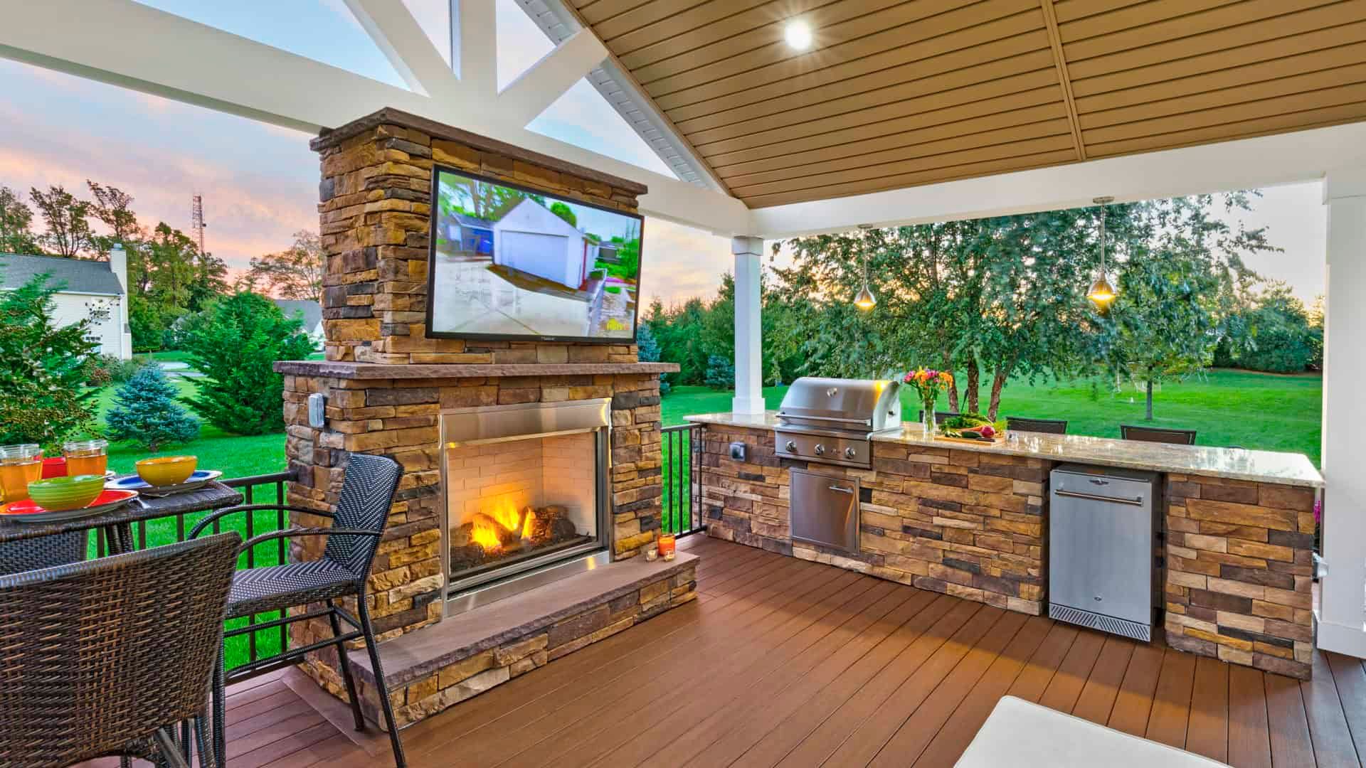 Covered patio with stone fireplace, mounted TV, outdoor seating, and built-in grill at sunset