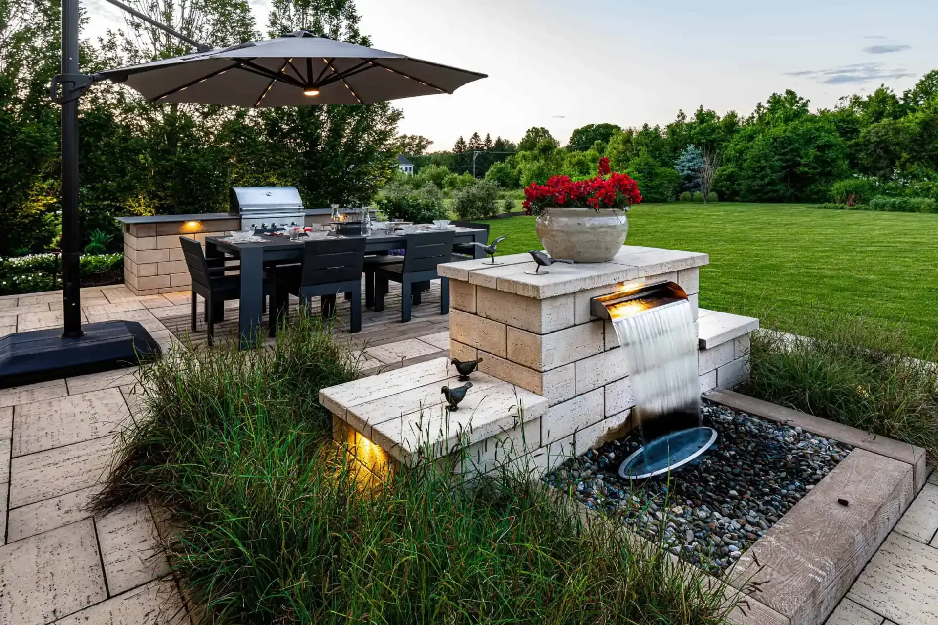 Backyard patio with dining table, umbrella, and stone fire feature beside a green lawn