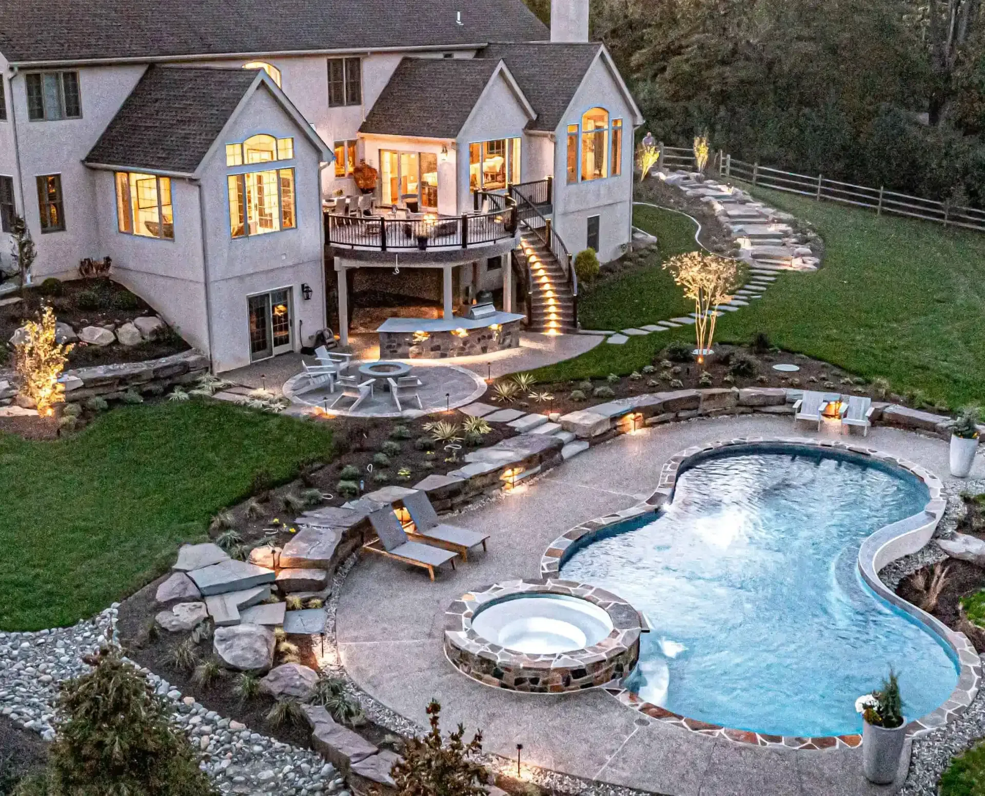 Luxurious backyard with a lit stone house, swimming pool, spa, deck, and landscaped garden at dusk