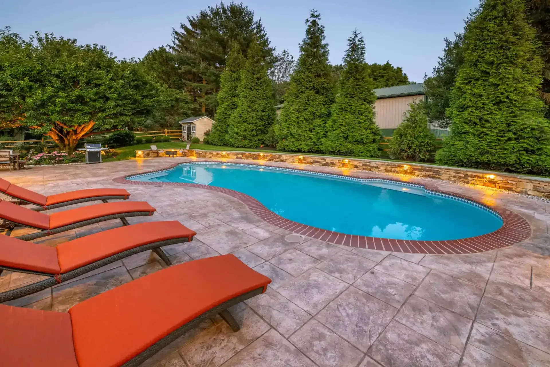 Backyard pool with orange lounge chairs, stone patio, and lush green trees surrounding it