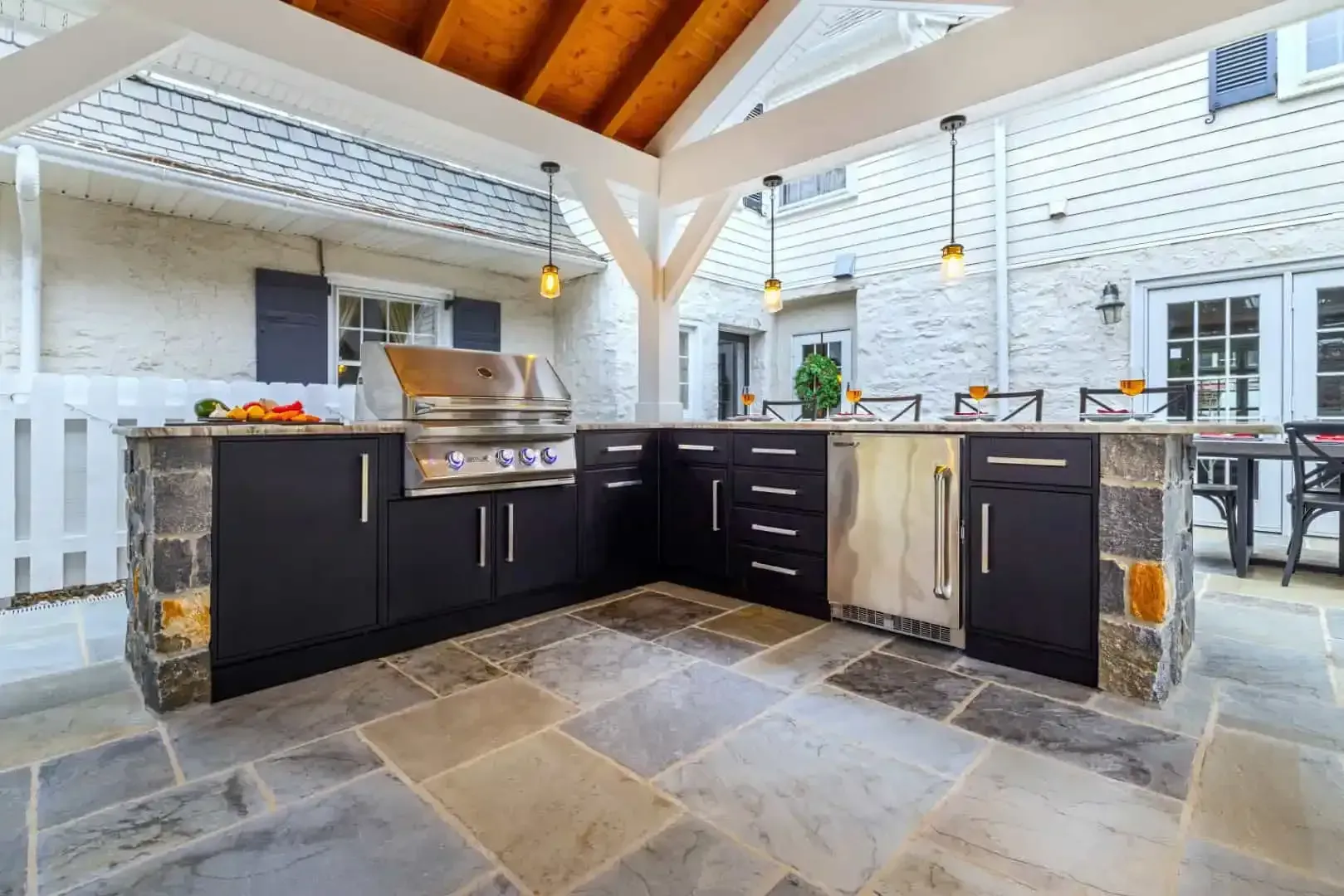 Outdoor kitchen with black cabinets, stainless grill, and stone countertops under a wood-beamed roof