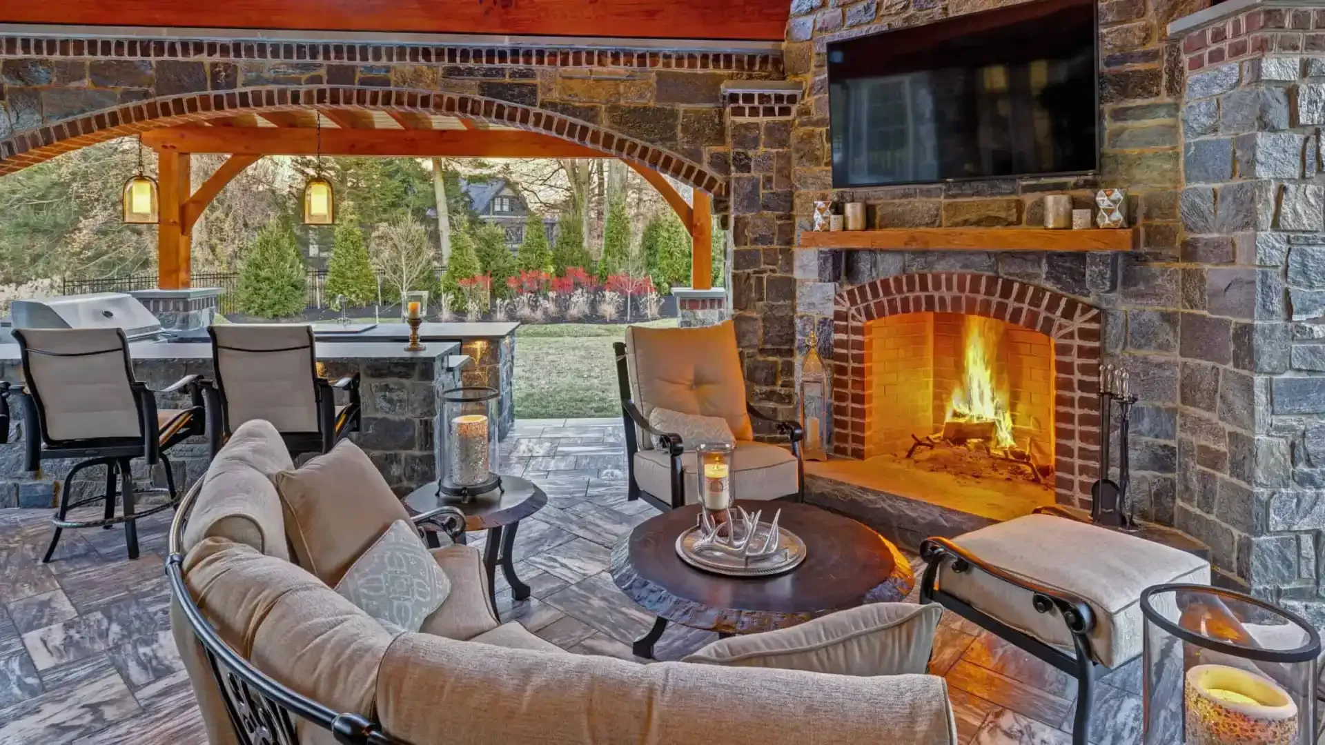 Cozy stone patio with lit fireplace, cushioned chairs, and outdoor dining area under a wooden pergola