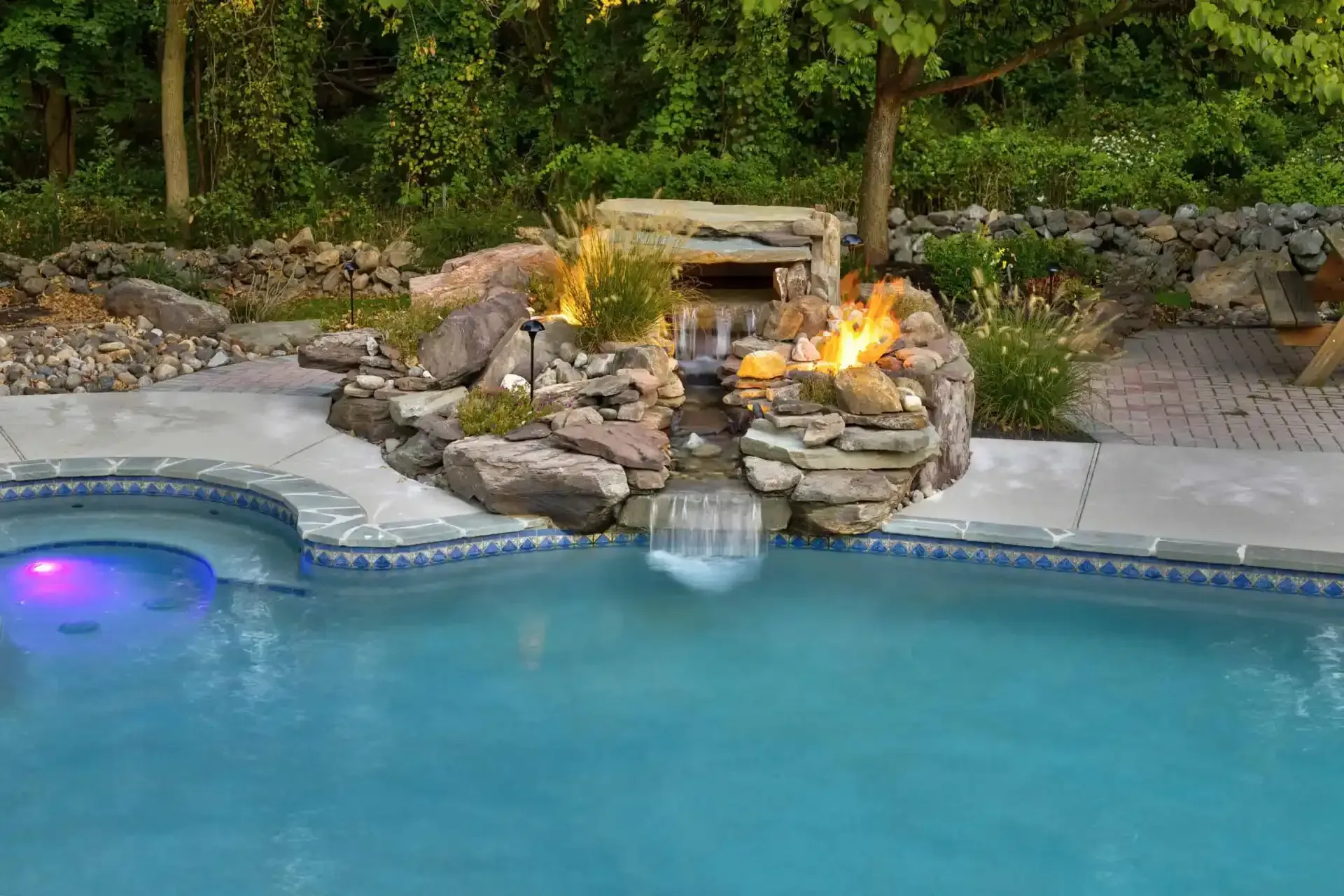 Backyard pool with stone waterfall, fire feature, and glowing spa at dusk