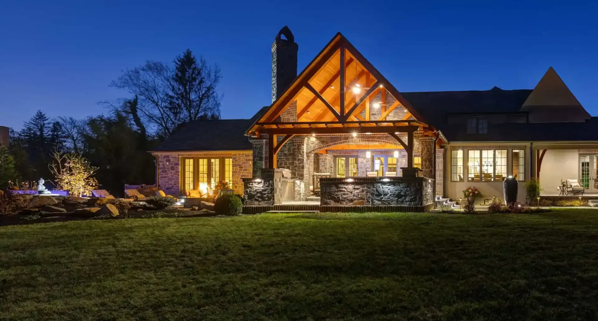 Illuminated stone house with a peaked timber entry at dusk, set behind a grassy lawn and trees.