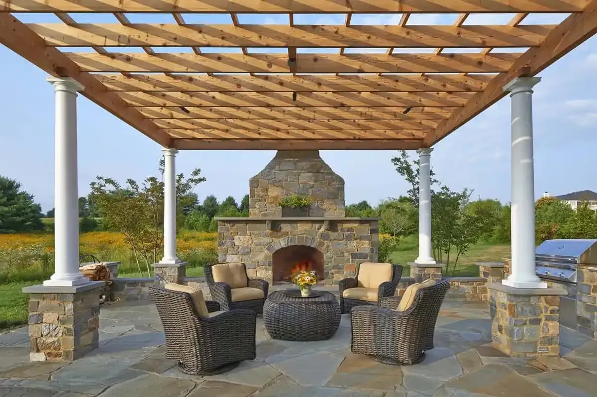Stone patio seating area with wicker chairs around a fire pit under a wood pergola, facing an outdoor fireplace