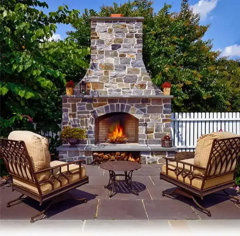 Backyard stone fireplace with glowing fire, flanked by two cushioned chairs on a patio.