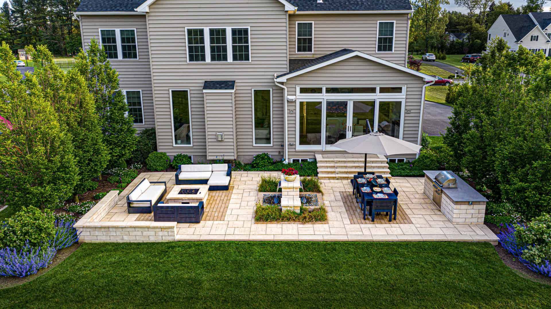 Modern backyard patio with white outdoor seating, dining set, and fire pit beside a beige house