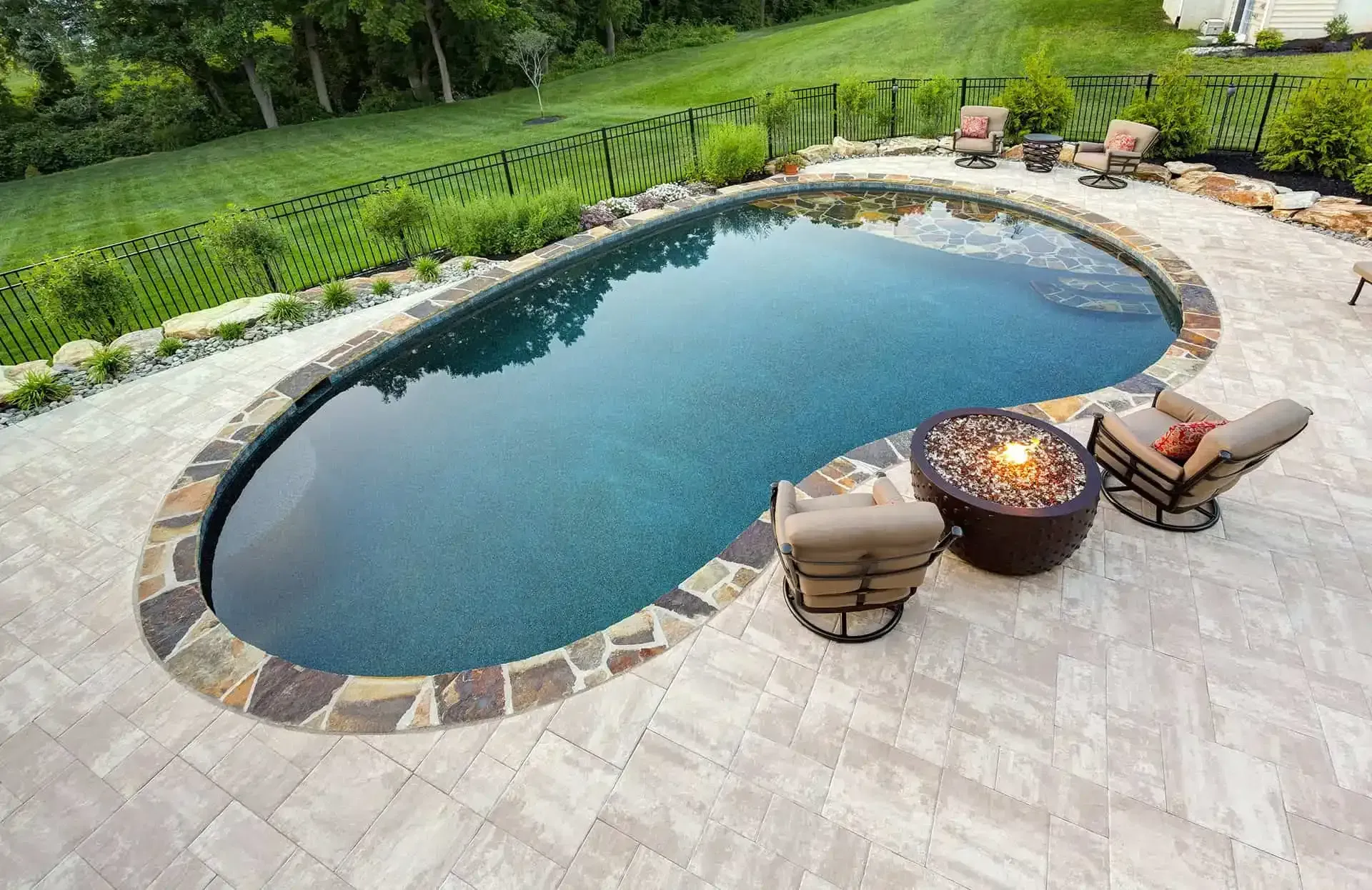Backyard oval swimming pool with stone patio, lounge chairs, and fire pit surrounded by lush greenery